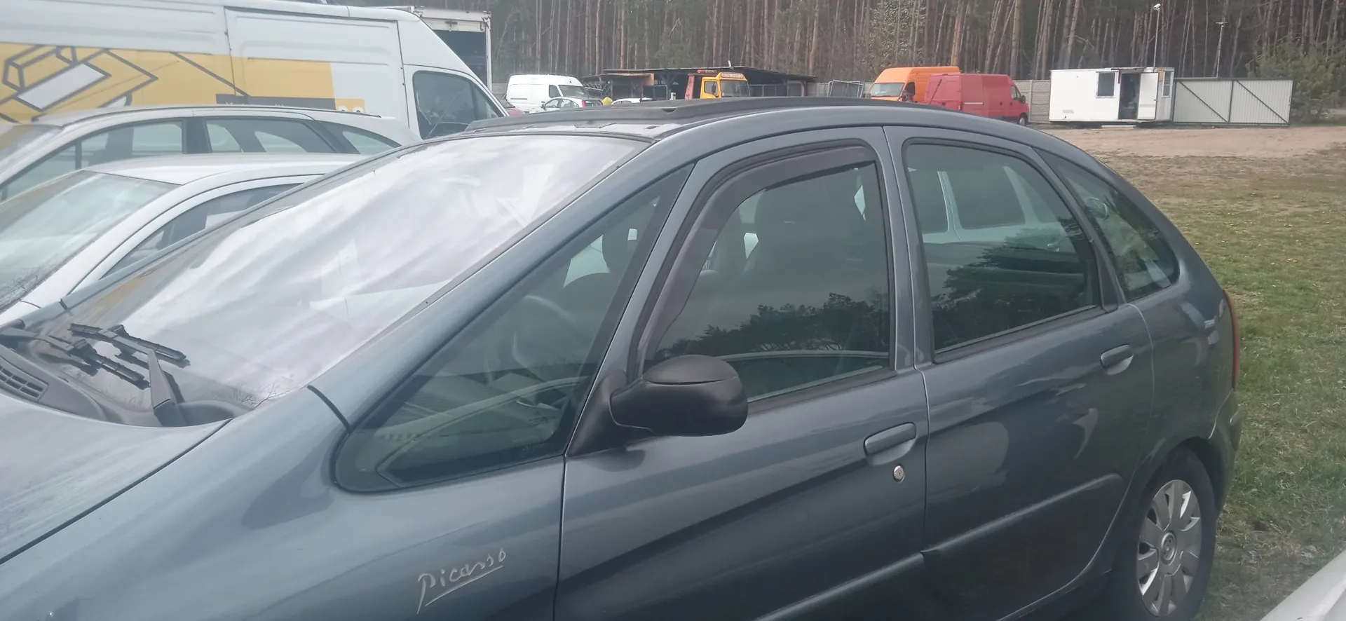 Szary samochód marki Citroën Picasso zaparkowany na zewnątrz. Widoczny jest bok auta z napisem 'Picasso' na przednim błotniku. W tle znajdują się inne pojazdy, w tym biały i pomarańczowy furgon, oraz las drzew.