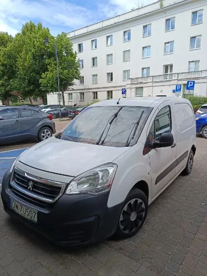 Jasnoszary samochód dostawczy marki Peugeot zaparkowany na kostce brukowej przed białym, wielopiętrowym budynkiem. Pojazd ma polską tablicę rejestracyjną o numerze DW 7U507. W tle widoczne są inne zaparkowane samochody, zielone drzewa oraz niebieskie znaki parkingowe na ścianie budynku.