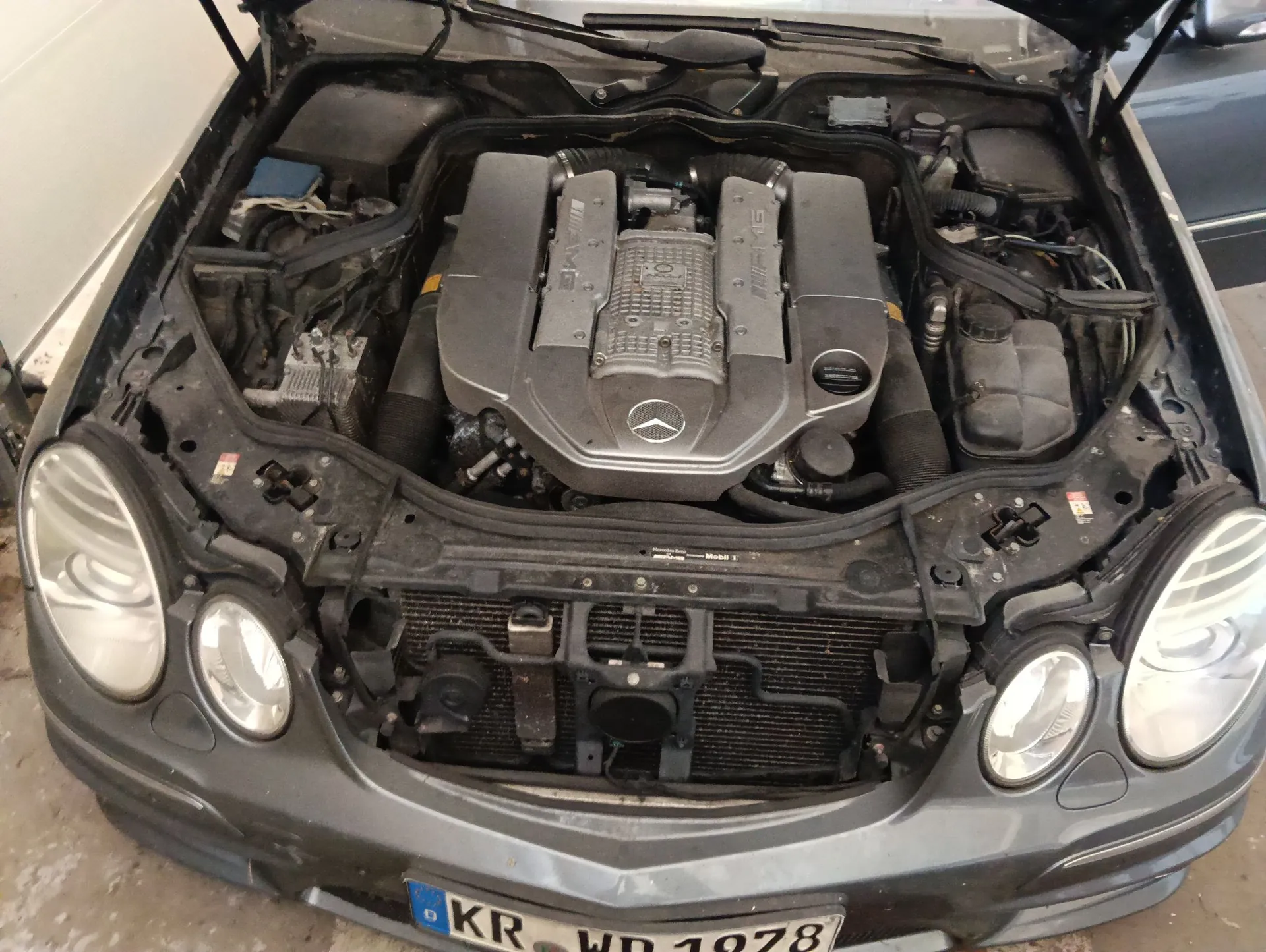 Widok z góry na otwartą maskę szarego samochodu marki Mercedes-Benz, ukazujący silnik V8 z charakterystycznymi osłonami w kolorze srebrnym z logo marki na środku. Widoczne są reflektory przednie, chłodnica oraz fragment tablicy rejestracyjnej o numerze KR WD 1978.