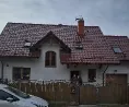 Jedno piętro dom jednorodzinny z białymi ścianami i spadzistym dachem pokrytym czerwoną dachówką. W dachu znajdują się okna połaciowe, a na szczycie komin. Przed domem znajduje się drewniany płot, za którym widoczne są dwie osoby. W lewym dolnym rogu zdjęcia widoczny jest fragment białego samochodu.