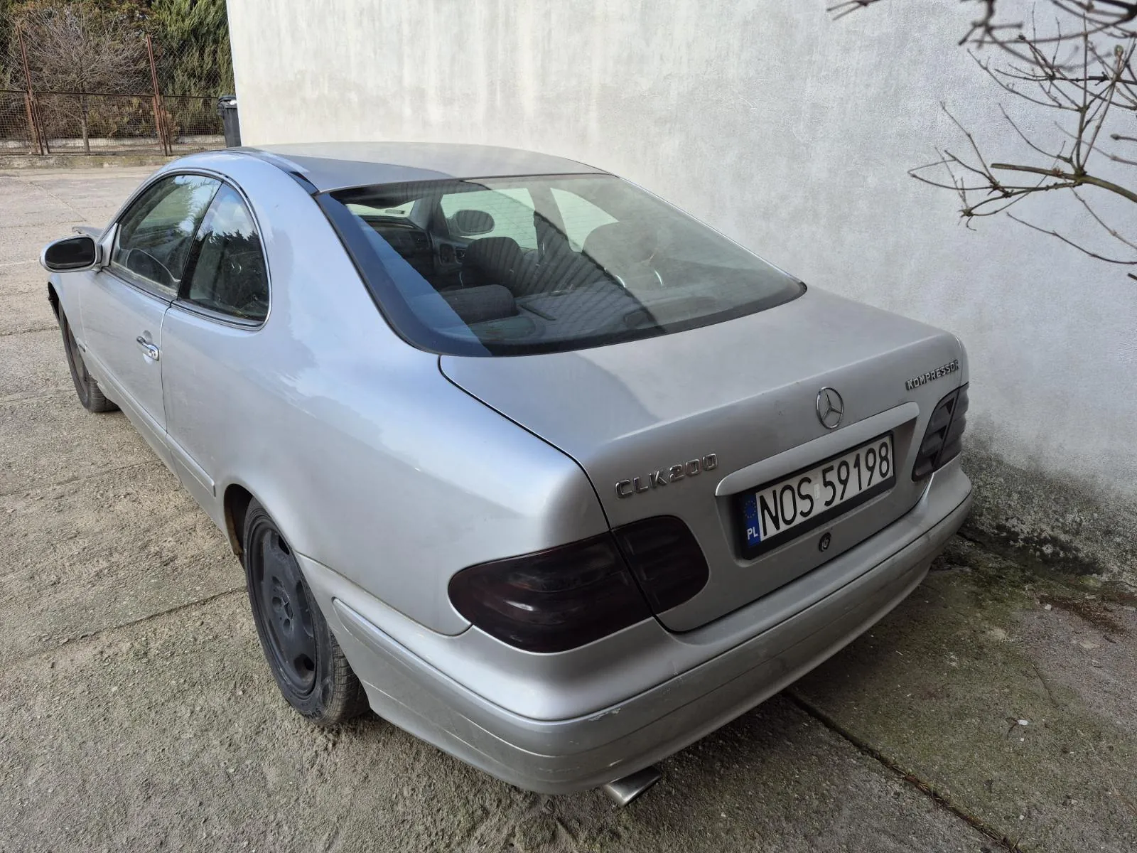 Srebrny samochód Mercedes-Benz CLK200 Kompressor zaparkowany na betonowej powierzchni obok szarej ściany. Widoczna tablica rejestracyjna z napisem NOS 59198 oraz oznaczeniem PL. Na bagażniku umieszczono emblemat Mercedes-Benz oraz napisy CLK200 i KOMPRESSOR. W tle widoczne są drzewa, metalowe ogrodzenie i sucha gałąź po prawej stronie. Wnętrze samochodu z widocznymi siedzeniami i kierownicą.