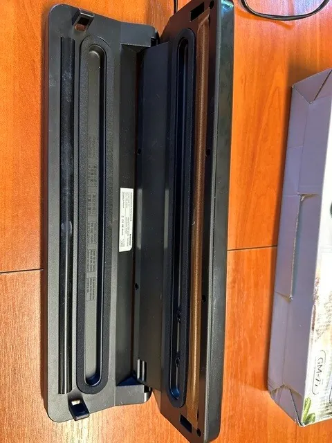 Zdjęcie przedstawiające dwa czarne, plastikowe elementy obudowy urządzenia elektronicznego leżące obok siebie na drewnianym blacie. Elementy mają podłużny kształt z wgłębieniami i technicznymi oznaczeniami na wewnętrznej stronie jednego z nich. Po prawej stronie widoczny jest fragment szarego kartonowego opakowania z napisem 'GM-7'.