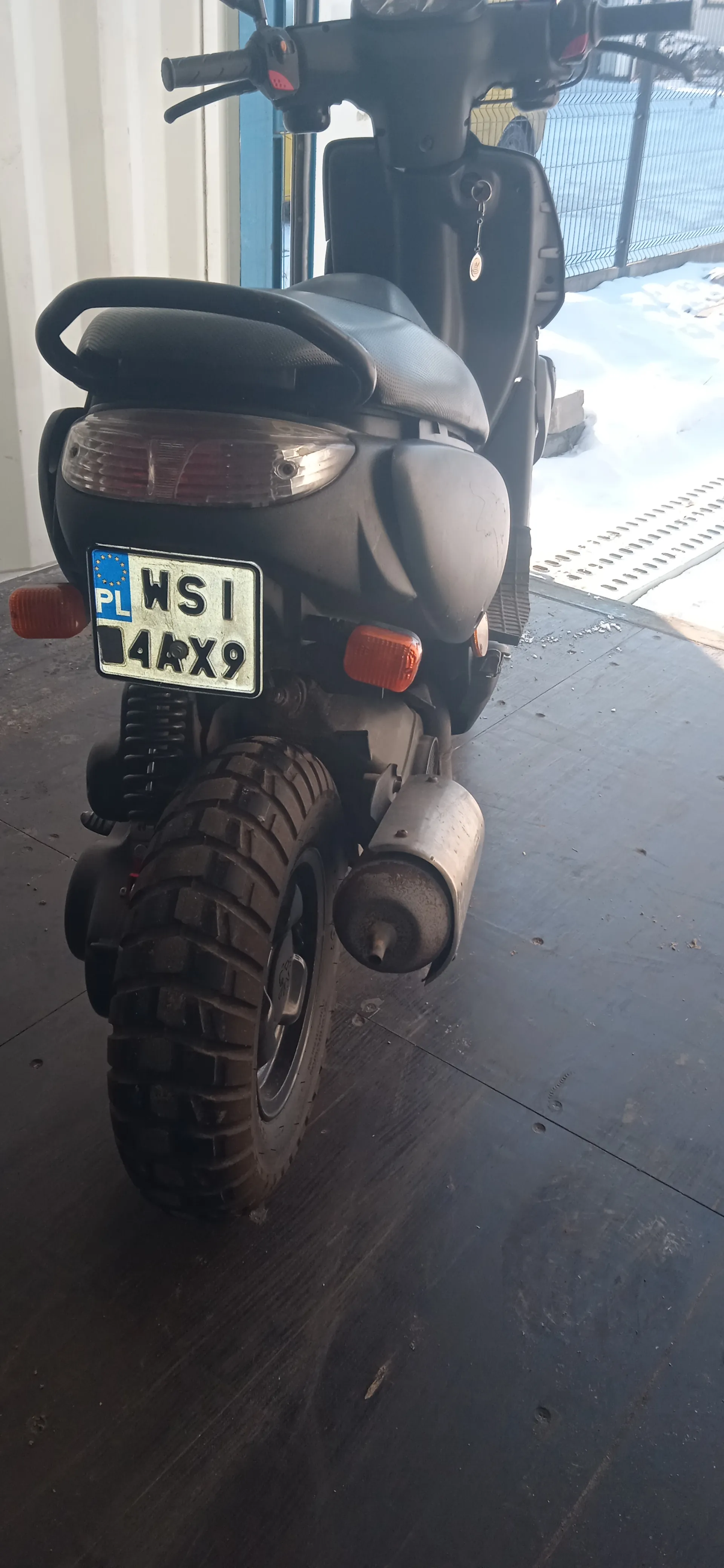 Czarny skuter widziany od tyłu z tablicą rejestracyjną PL WSI 4AX9. Skuter stoi na ciemnej podłodze, w tle widać śnieg i ogrodzenie. Widoczne są kierownica, siedzenie, tylna opona oraz wydech.