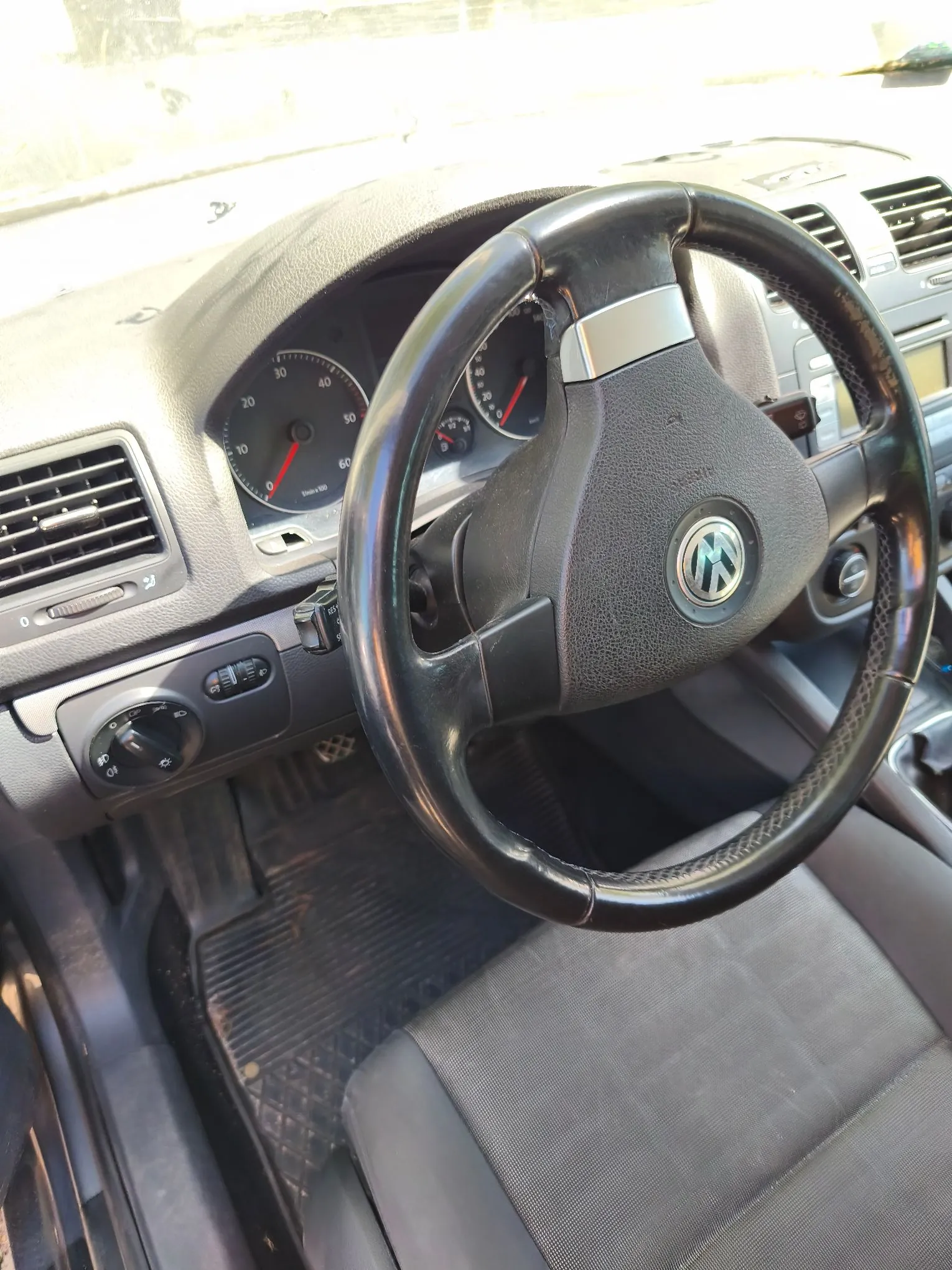 Wnętrze samochodu marki Volkswagen widziane z perspektywy kierowcy. W centrum znajduje się czarna, skórzana kierownica z logo VW. W tle widoczna jest deska rozdzielcza z analogowymi wskaźnikami prędkości i obrotów, kratki nawiewów oraz panel sterowania klimatyzacją. W dolnej części zdjęcia widoczny jest fragment czarnego fotela oraz gumowa mata na podłodze.