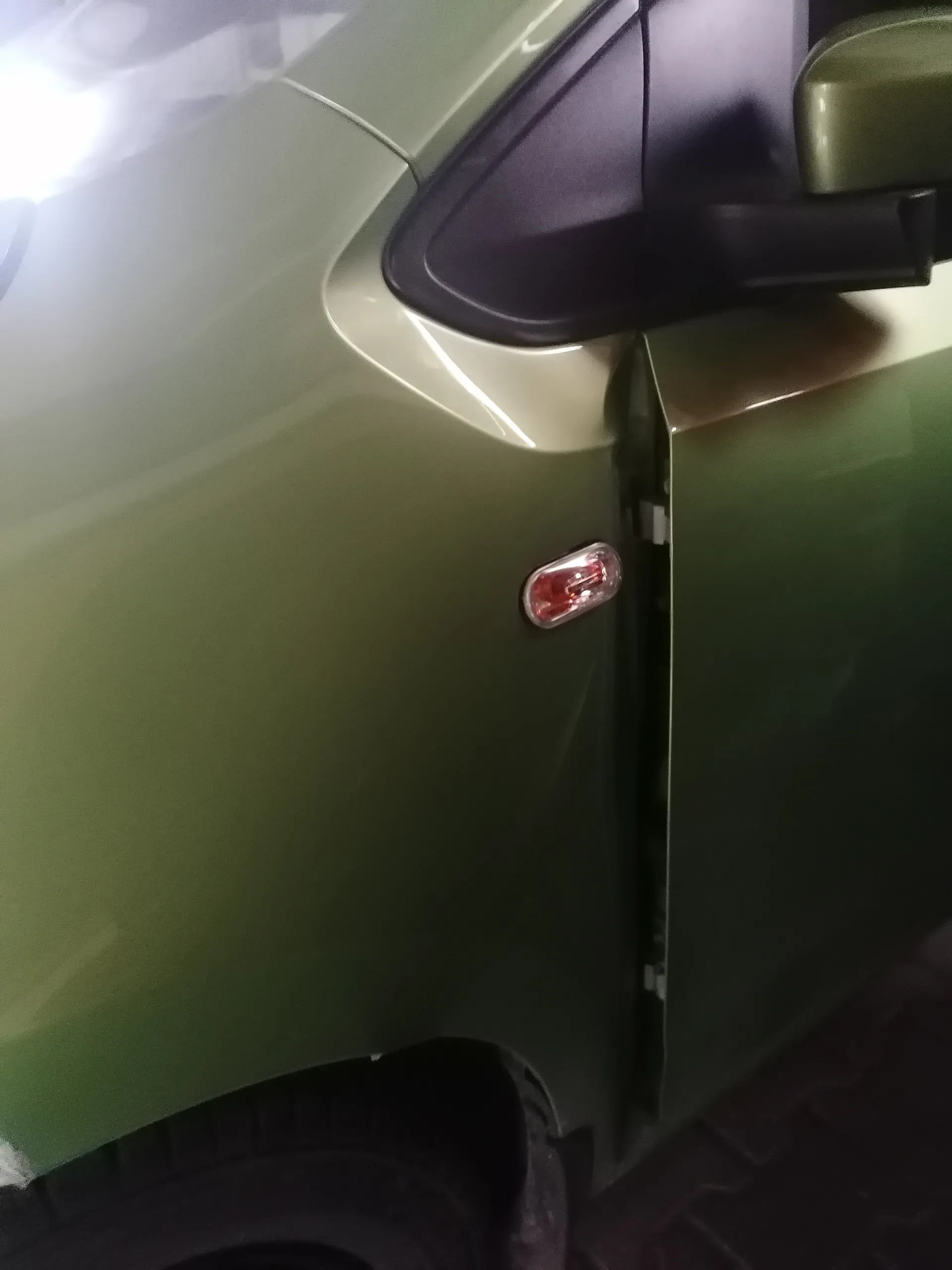 Zbliżenie na zielony samochód. Widoczna część przedniego błotnika, czarno-zielone lusterko boczne, czerwone światło sygnalizacyjne na błotniku oraz fragment koła z oponą. Tło stanowi ciemna, brukowana powierzchnia.