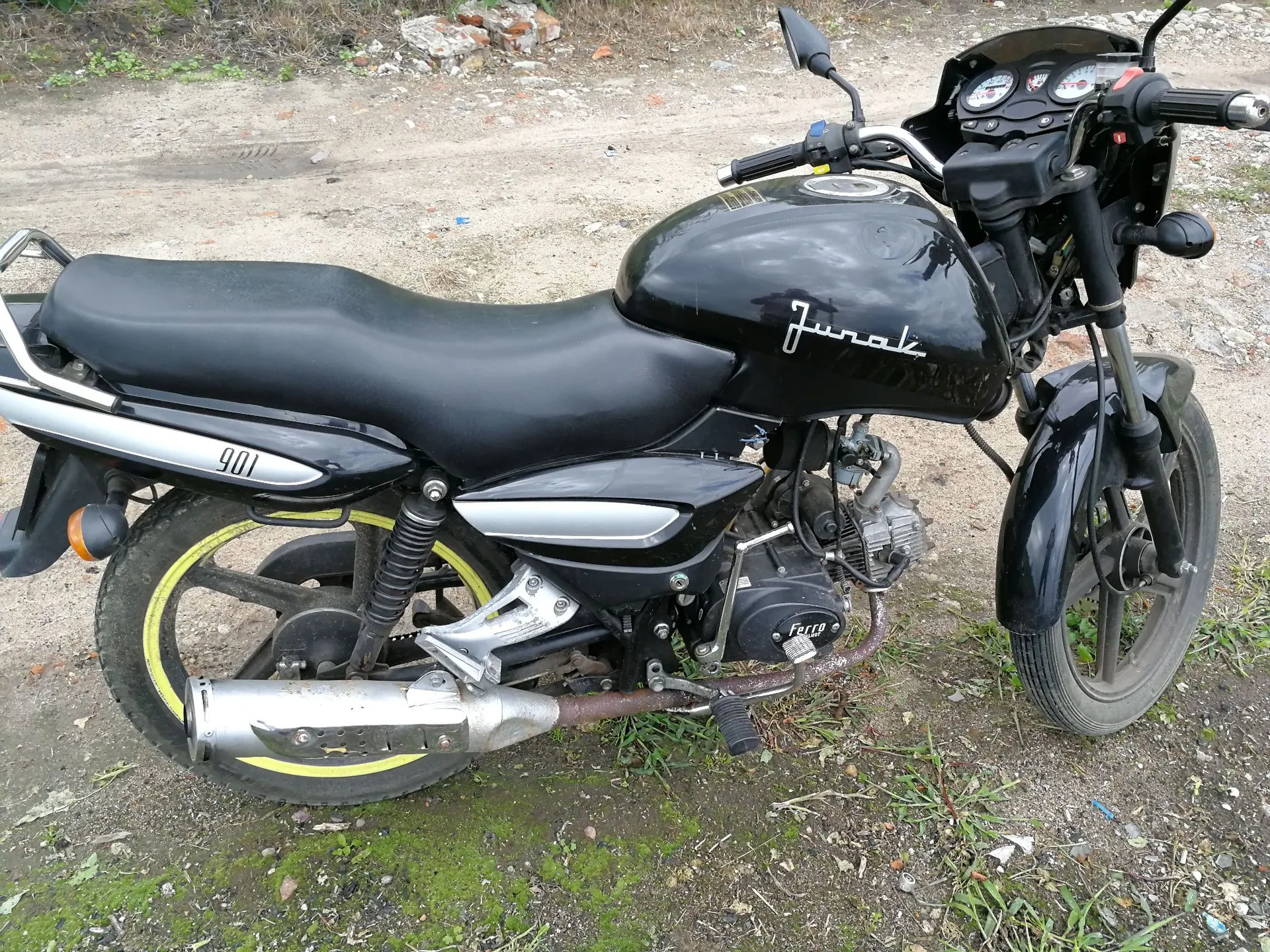 Czarny motocykl zaparkowany na piaszczystym i trawiastym podłożu. Na baku paliwa widnieje biały napis 'Jurak', a na bocznej owiewce liczba '100'. Pojazd posiada srebrny wydech, czarne felgi z żółtą obwódką na przednim kole oraz czarne siedzenie.