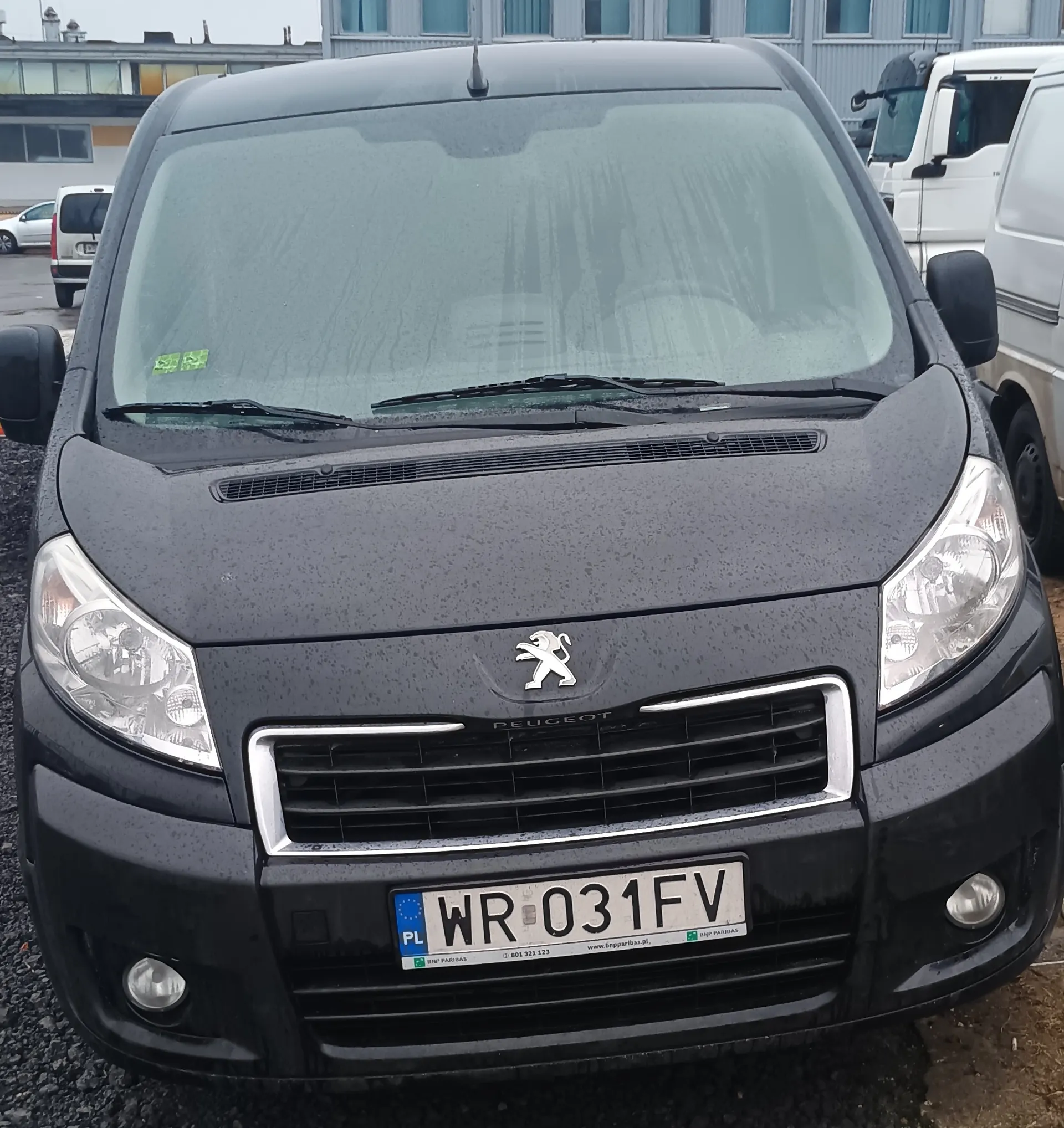 Czarny samochód dostawczy marki Peugeot z przodu, z widocznym logo lwa na masce i tablicą rejestracyjną WR 031FV z polskim oznaczeniem PL. Na przedniej szybie znajdują się krople deszczu oraz zielony naklejka. W tle widoczne są inne pojazdy (biały furgon i ciężarówka) oraz budynki z oknami. Samochód stoi na podłożu z żwiru.