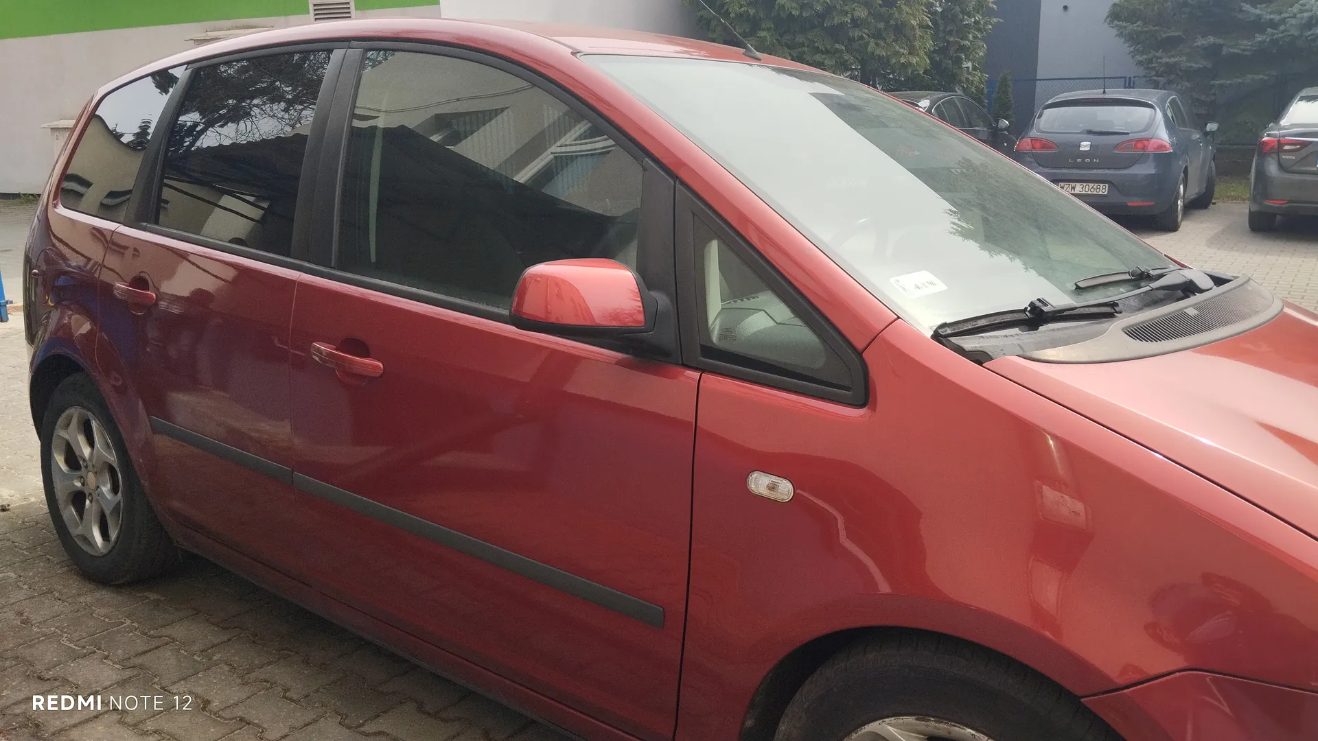 Czerwony samochód osobowy typu hatchback zaparkowany na brukowanej powierzchni. W tle widoczne są inne pojazdy: niebieski samochód z tablicą rejestracyjną WZW 30688 oraz szary samochód. W tle również budynki z zielonym pasem i drzewa. W lewym dolnym rogu obrazu znajduje się napis "REDMI NOTE 12".