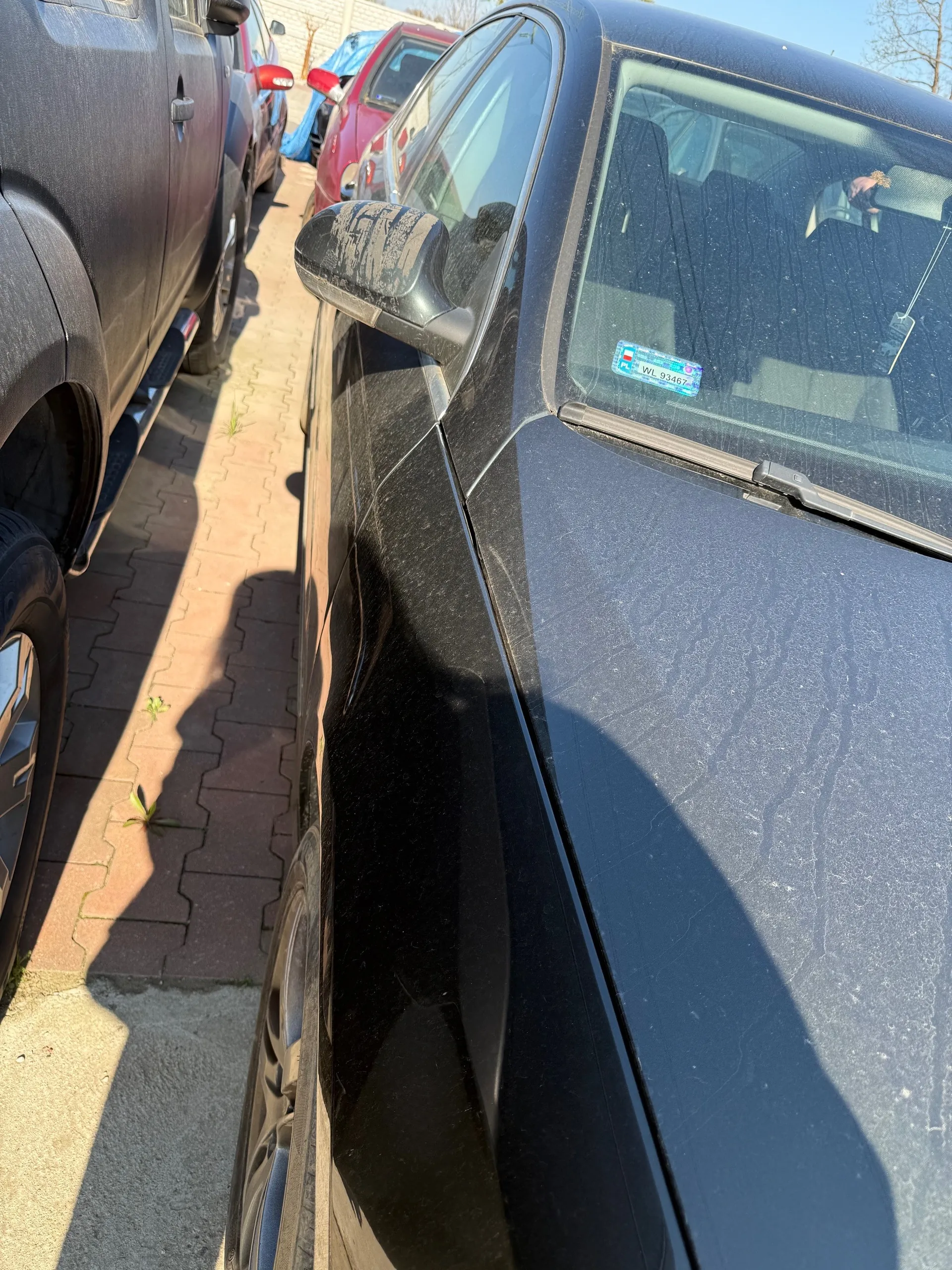 Czarny samochód zaparkowany na brukowanej powierzchni, widok z boku. Na przedniej szybie naklejka z niebieskim tłem i białym tekstem: WIL 93467. Obok czarnego samochodu widoczne są inne pojazdy: ciemnoszary SUV po lewej stronie oraz czerwony i niebieski samochód w tle. Na ziemi płytki brukowe z pojedynczymi roślinami. Cień osoby fotografującej widoczny na powierzchni parkingu.