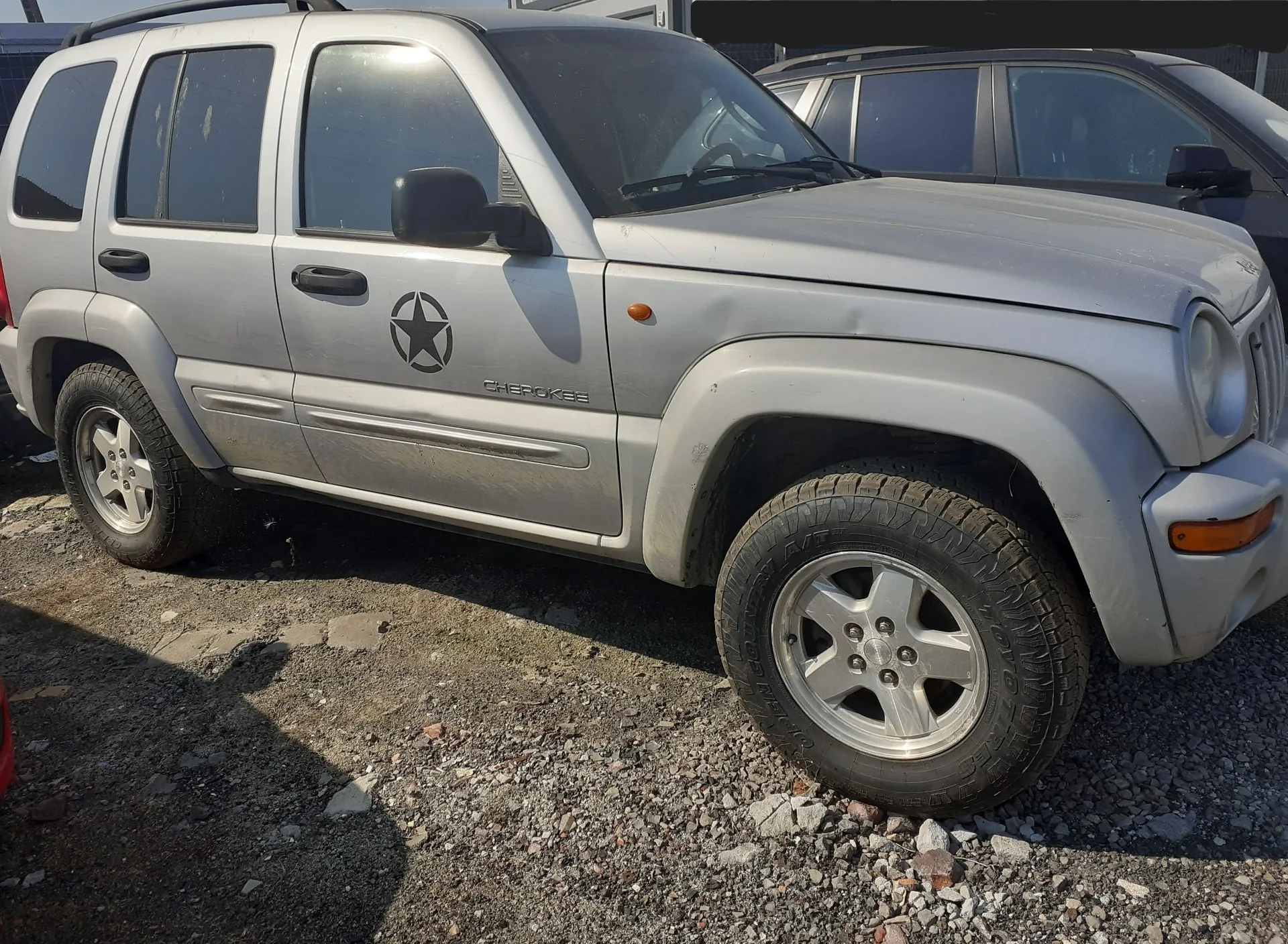 Srebrny samochód terenowy Jeep Cherokee zaparkowany na żwirowym podłożu. Na przednich drzwiach kierowcy znajduje się czarna naklejka przedstawiająca gwiazdę w okręgu oraz napis 'CHEROKEE'. Pojazd ma widoczne wgniecenie na przednim błotniku nad nadkolem. W tle widoczny jest fragment innego, ciemnego samochodu.