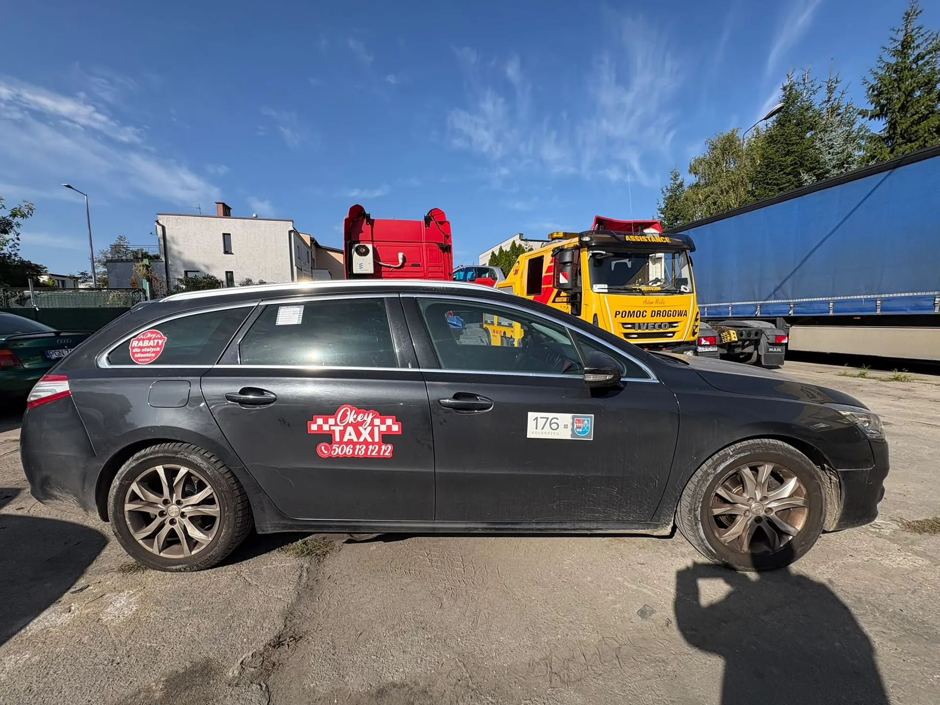 Szary samochód osobowy typu kombi zaparkowany bokiem do obiektywu. Na drzwiach kierowcy znajduje się czerwono-biała naklejka z napisem 'Okey TAXI' oraz numerem telefonu '506 13 12 12'. Na tylnej szybie widnieje okrągła naklejka z napisem 'RABATY'. W tle widoczny jest żółty samochód pomocy drogowej z napisem 'POMOC DROGOWA IVECO', czerwony pojazd specjalistyczny oraz niebieska plandeka ciężarówki. W oddali znajduje się biały budynek mieszkalny pod niebieskim niebem z lekkimi chmurami.