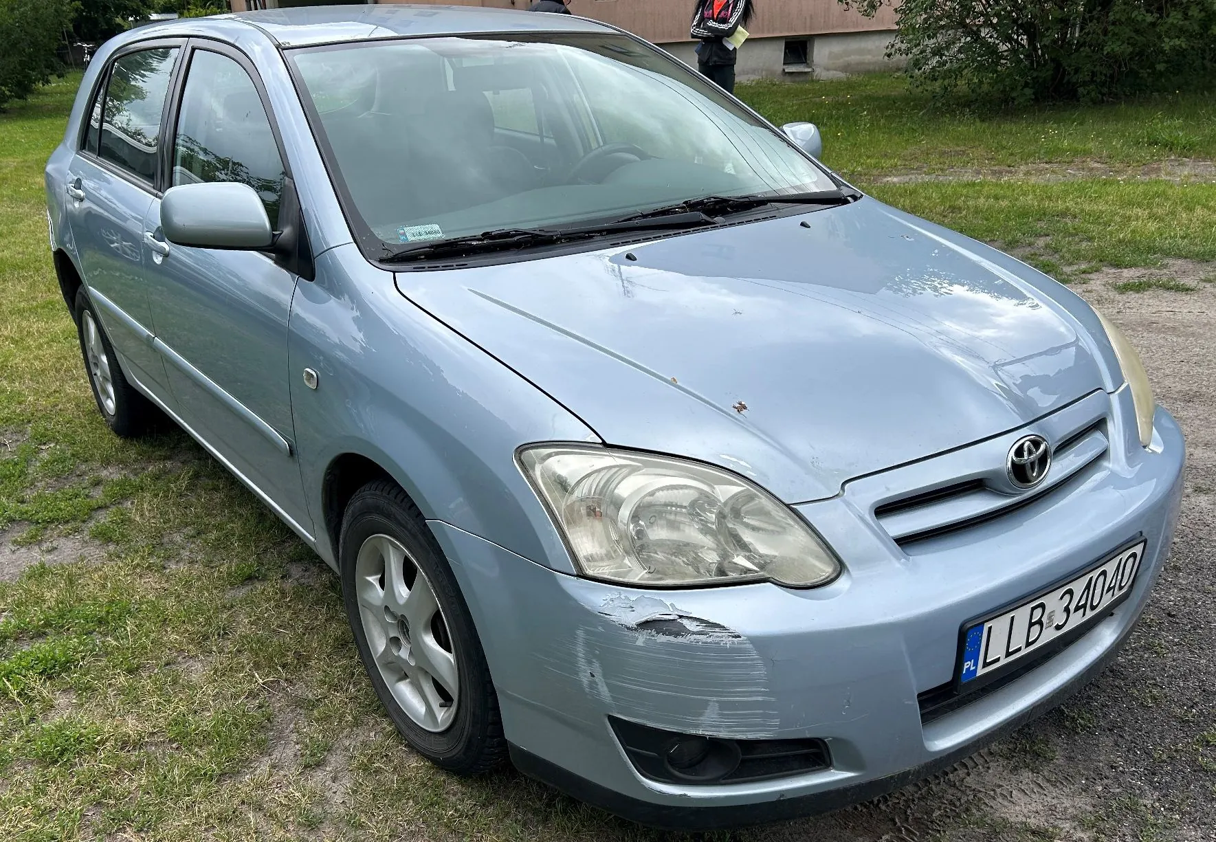 Jasnoniebieski samochód osobowy marki Toyota zaparkowany na trawiastym podłożu. Pojazd jest widoczny z przodu i z boku. Na przednim zderzaku, po stronie pasażera, widoczne są zarysowania i uszkodzenia lakieru. Tablica rejestracyjna ma numer LLB 34040.