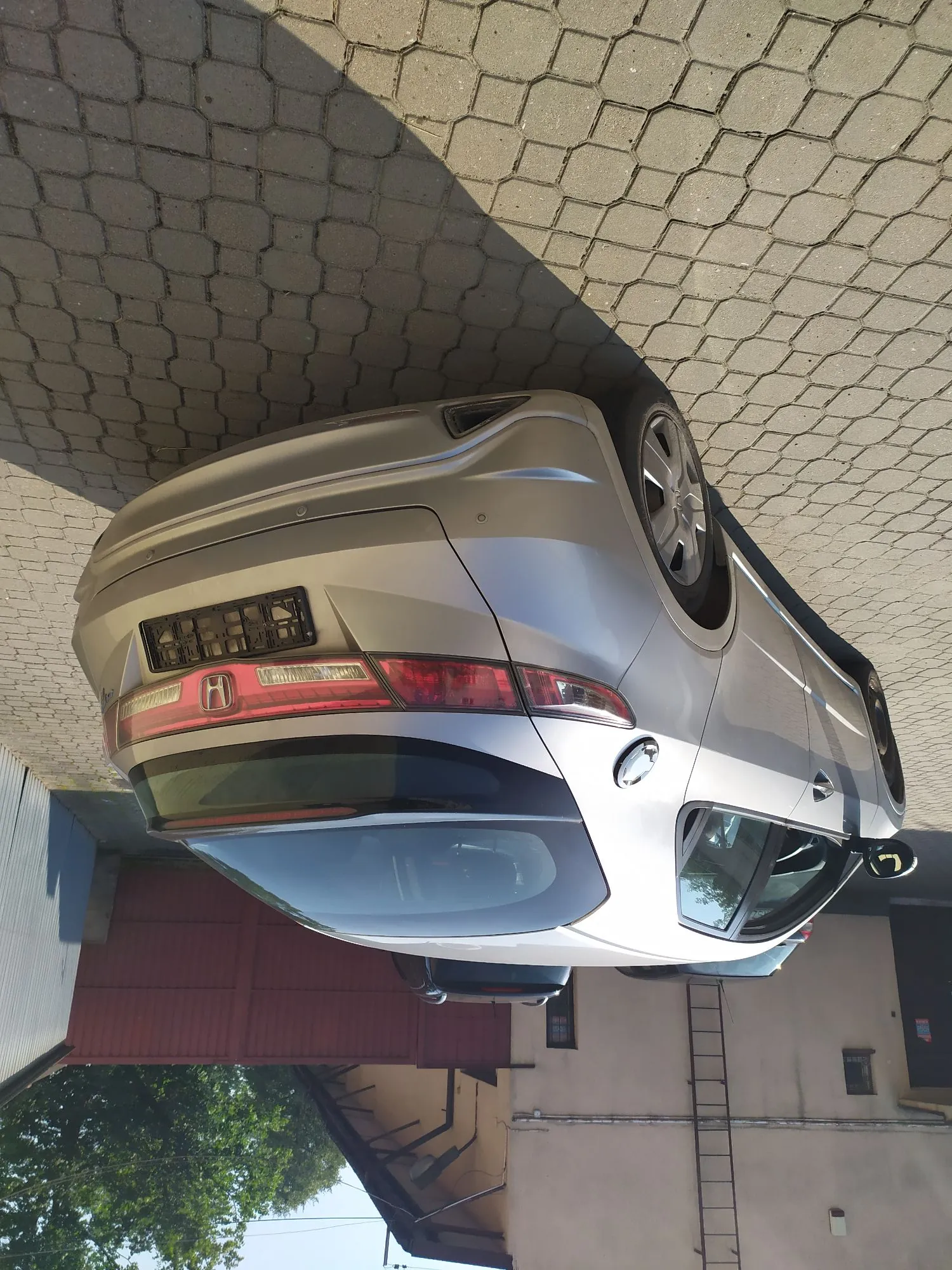 Srebrny samochód marki Honda zaparkowany na kostce brukowej. Zdjęcie przedstawia tył i bok pojazdu. W tle widoczny jest budynek z czerwoną bramą oraz fragmenty zieleni.
