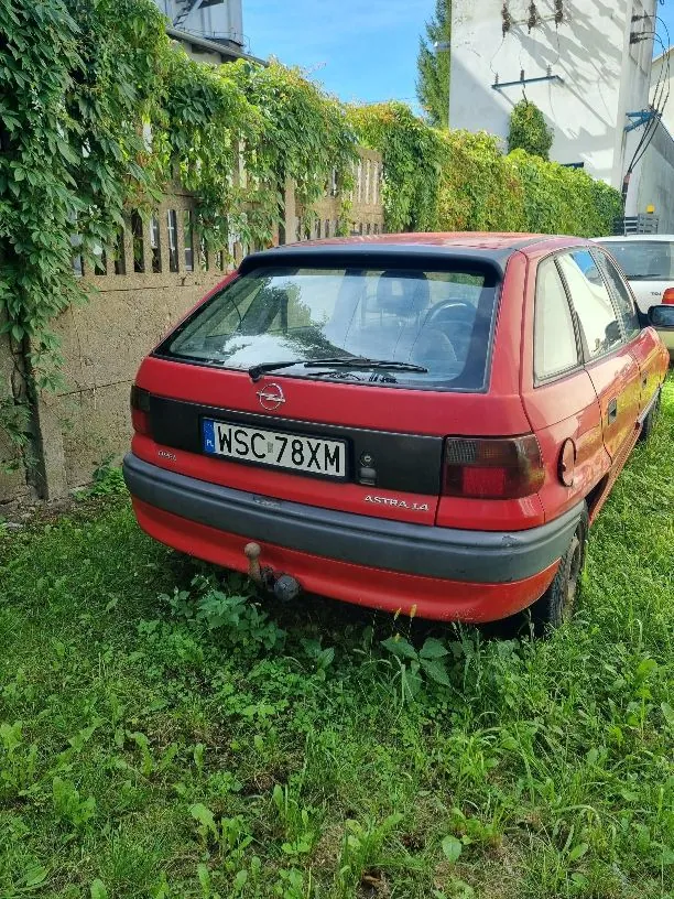 Tylny widok czerwonego samochodu marki Opel Astra 1.4 zaparkowanego na trawie. Pojazd posiada polską tablicę rejestracyjną o numerze WSC 78XM oraz zamontowany hak holowniczy. W tle widoczny jest betonowy płot porośnięty zielonymi roślinami oraz fragment białego budynku.
