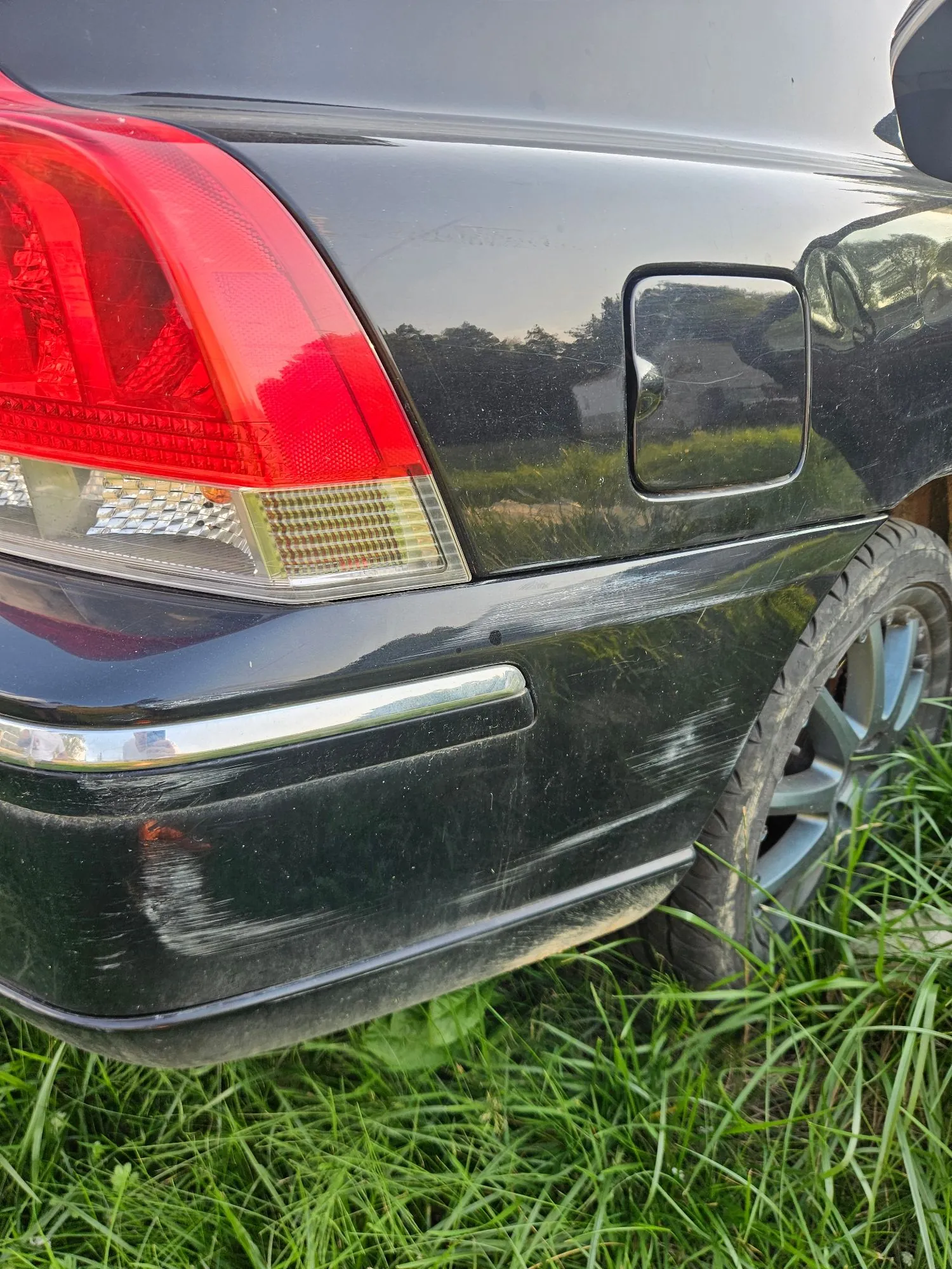 Zbliżenie na tylny lewy narożnik czarnego samochodu. Widoczna jest czerwona lampa tylna oraz uszkodzenia w postaci licznych zarysowań i otarć na lakierze zderzaka i błotnika w okolicach klapki wlewu paliwa. W kadrze znajduje się również fragment koła z felgą aluminiową, a samochód stoi na zielonej trawie.