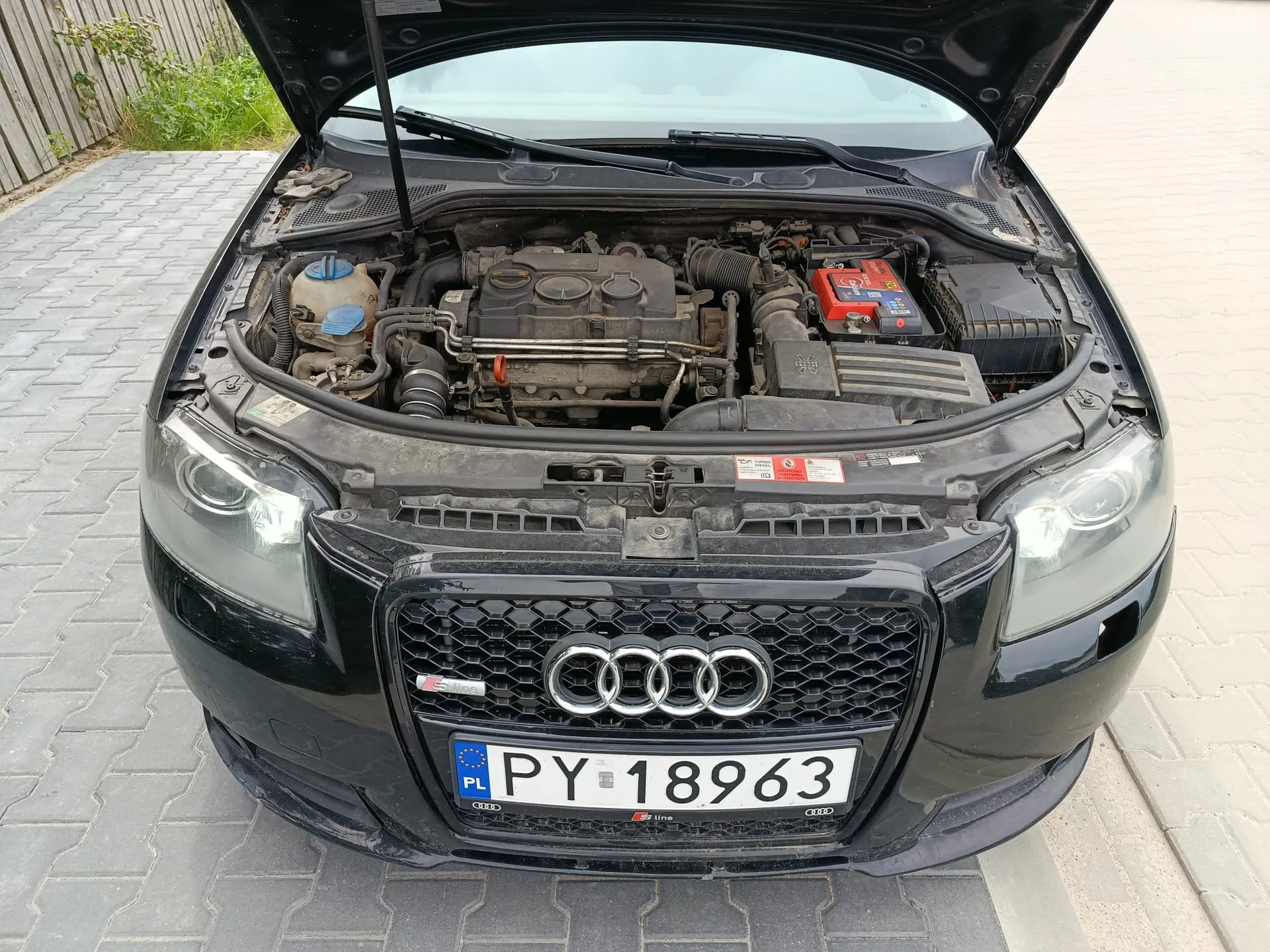 Czarny samochód marki Audi z otwartą maską, ukazującą silnik i komponenty komory silnika. Na przodzie pojazdu widoczna jest charakterystyczna atrapa chłodnicy z logo czterech pierścieni oraz polska tablica rejestracyjna o numerze PY 18963.