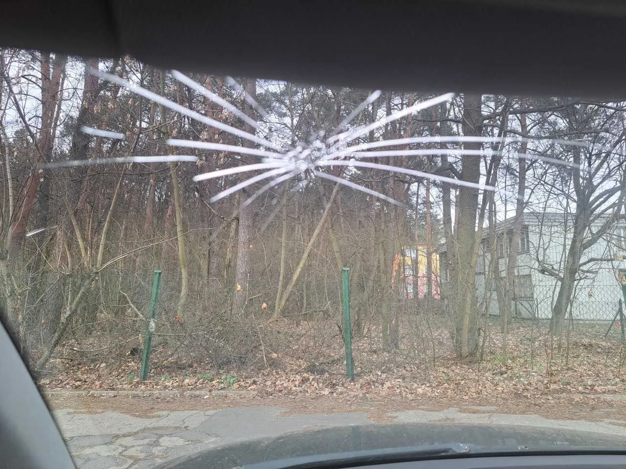 Widok z wnętrza samochodu przez przednią szybę z widocznym pęknięciem w kształcie pajęczyny. Na zewnątrz drzewa bez liści, zielone słupki ogrodzenia, domy w tle oraz ziemia pokryta suchymi liśćmi. Niebo pochmurne.