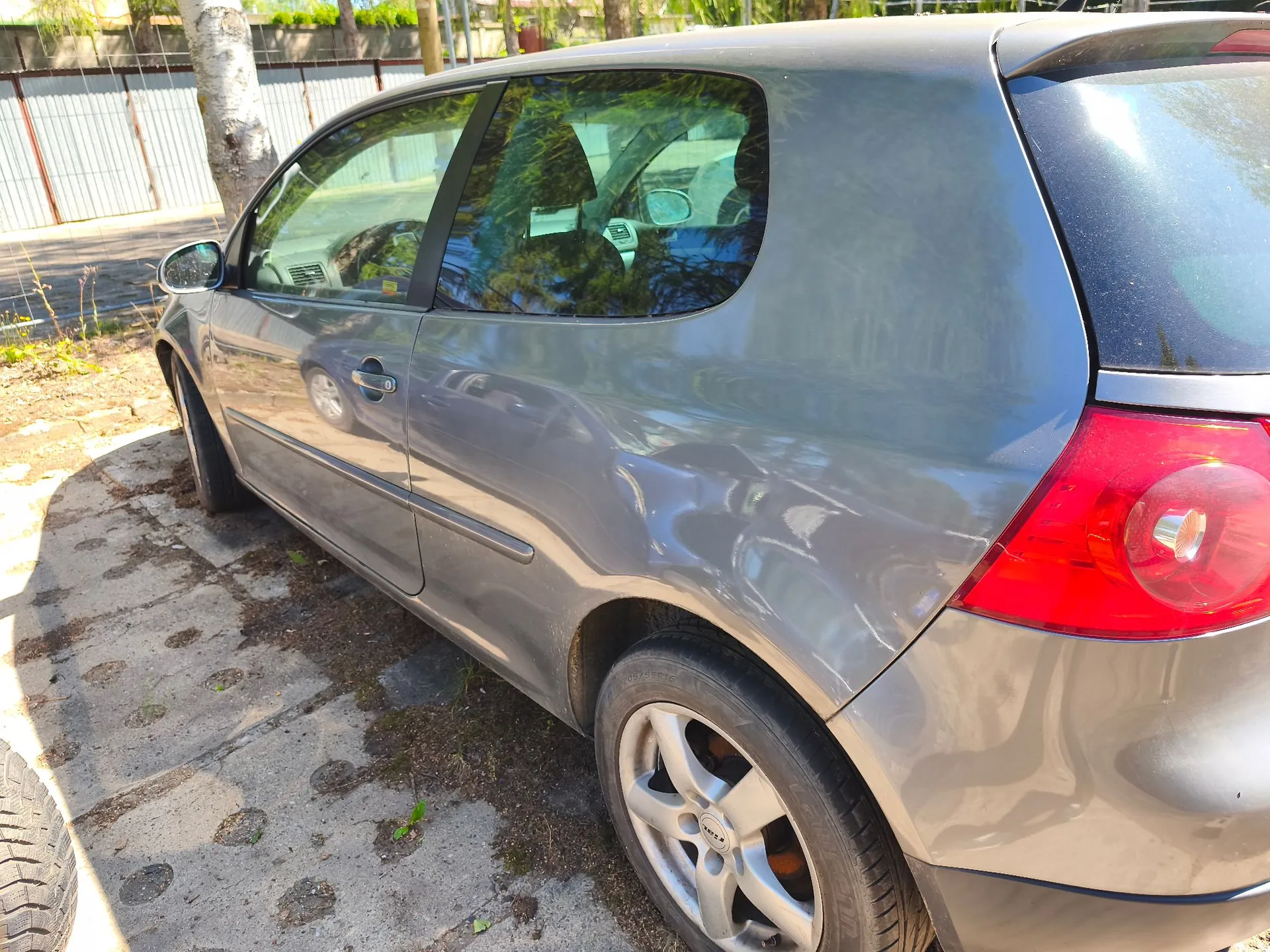 Szary samochód osobowy typu hatchback zaparkowany na betonowym podłożu. Zdjęcie przedstawia prawy bok pojazdu od tyłu, ukazując tylną lampę, koło z felgą aluminiową oraz drzwi pasażera. W tle widoczny jest biały płot i drzewa.