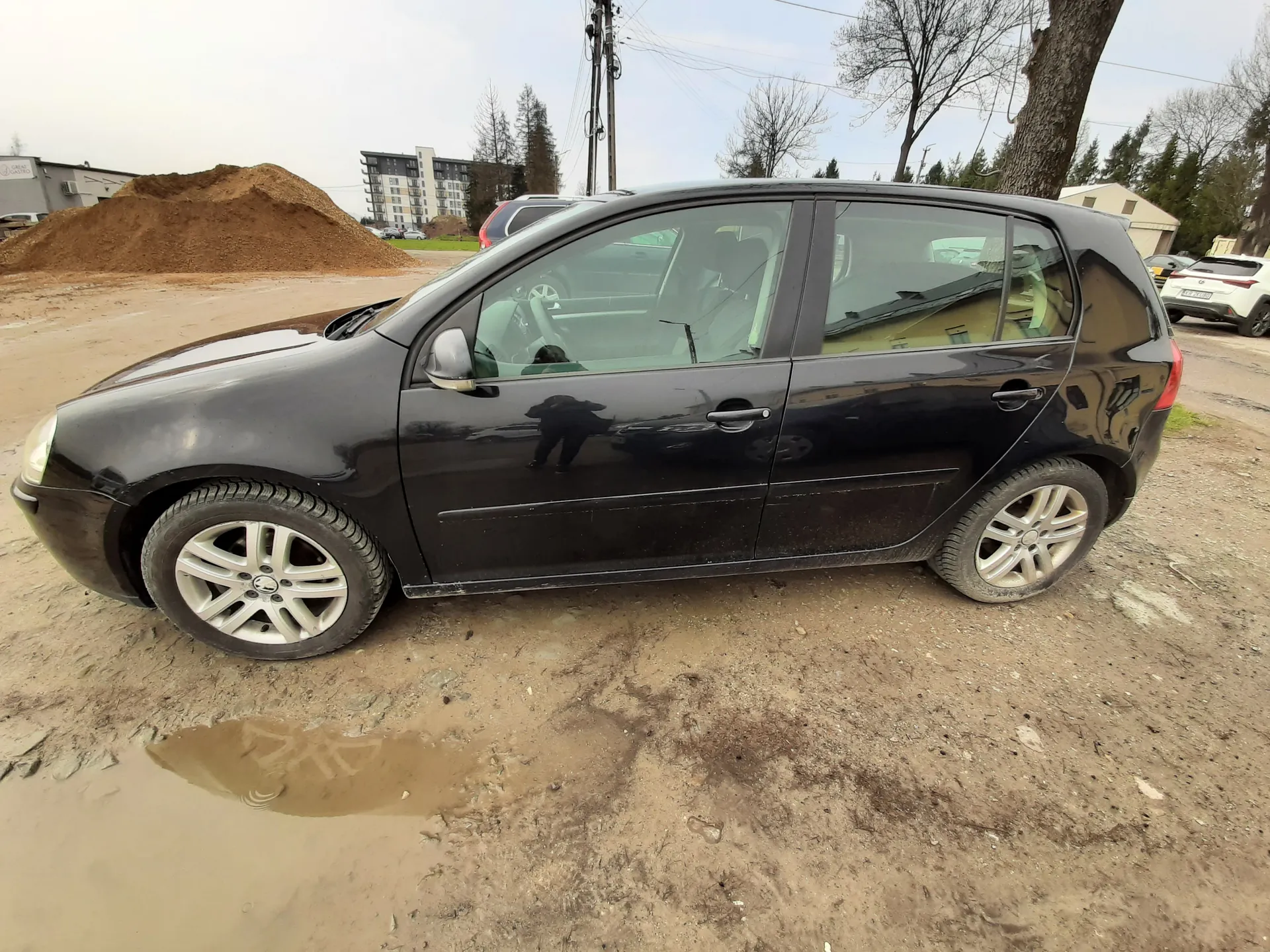 Czarny samochód osobowy typu hatchback zaparkowany na brudnej, piaszczystej powierzchni z kałużami. W tle widoczna duża kopca ziemi, budynki mieszkalne, drzewa bez liści, słup energetyczny oraz inne zaparkowane pojazdy. Na pierwszym planie odbicie osoby w drzwiach samochodu.