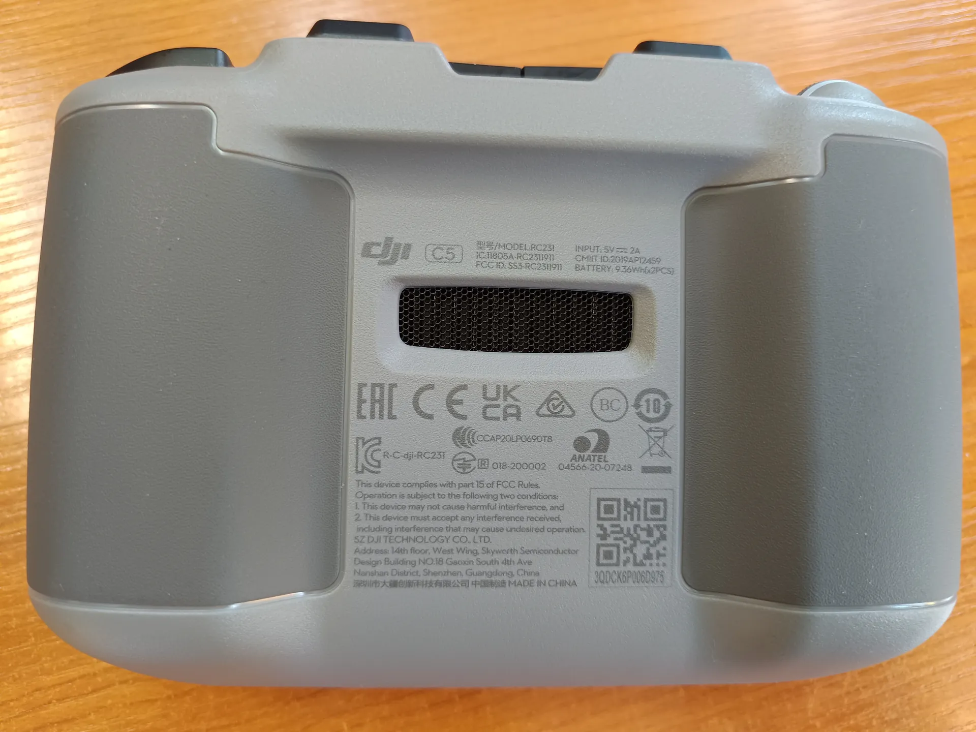 Szare urządzenie elektroniczne z pionowo umieszczonymi oznaczeniami: logo DJI, oznaczenia certyfikacyjne EAC, CE, UKCA, numer modelu RC231, dane wejściowe (5V --- 2A), informacje o baterii (9.26Wh), kod QR oraz tekst w języku chińskim i angielskim. Na powierzchni widoczne są również symbole certyfikacji, adres producenta w Shenzhen, Chiny oraz napis 'MADE IN CHINA'. Urządzenie leży na drewnianym tle.