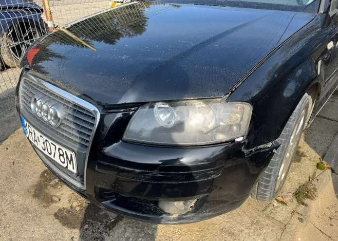 Czarny samochód marki Audi z przodu, widoczna maska, reflektory, chromowana atrapa chłodnicy z logo Audi oraz tablica rejestracyjna KRA-3078M. Samochód stoi na betonowej powierzchni, w tle siatka ogrodzeniowa i fragment innych pojazdów. Na masce widoczne są ślady brudu i uszkodzenia na przednim zderzaku po prawej stronie.