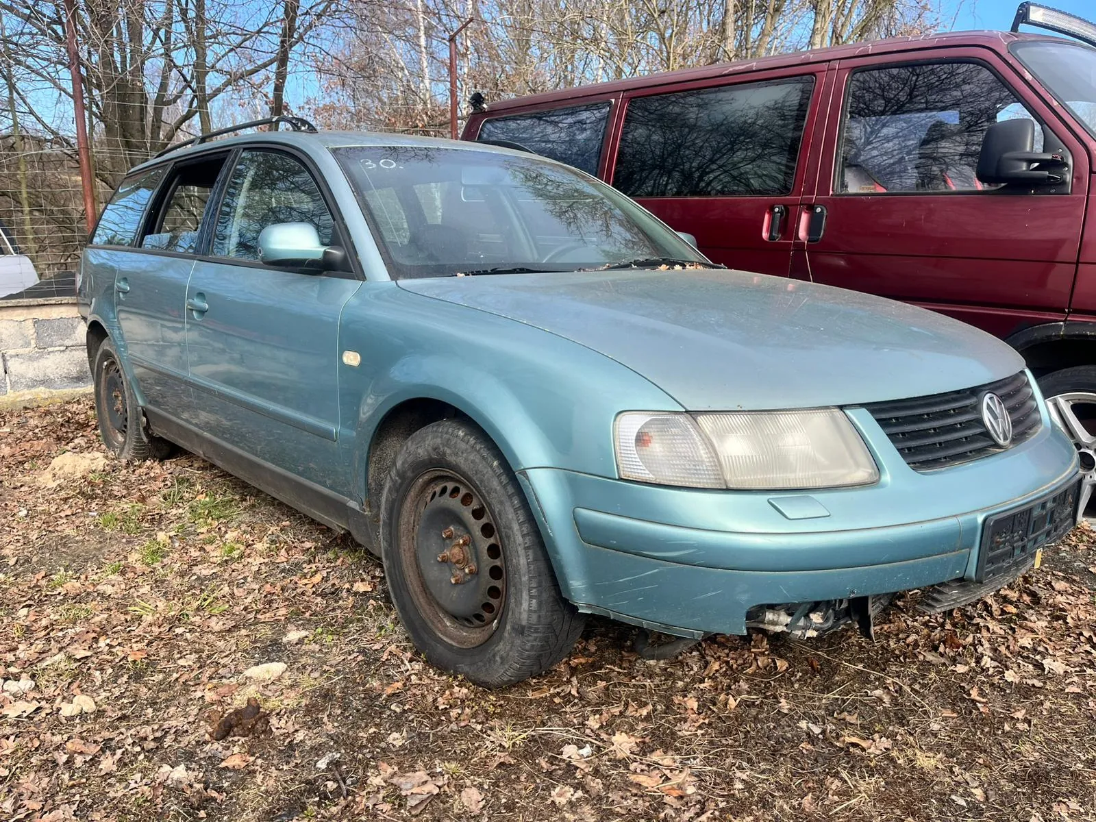 Niebieski samochód osobowy typu kombi marki Volkswagen z uszkodzonym przodem i brakiem przedniego zderzaka. Na przedniej szybie widoczne oznaczenie '30.'. Samochód stoi na podłożu z suchą trawą i opadłymi liśćmi, z zardzewiałymi felgami. W tle czerwony van, drzewa bez liści, fragment ogrodzenia i kamienna ściana. Niebo jest jasne, pogoda słoneczna.