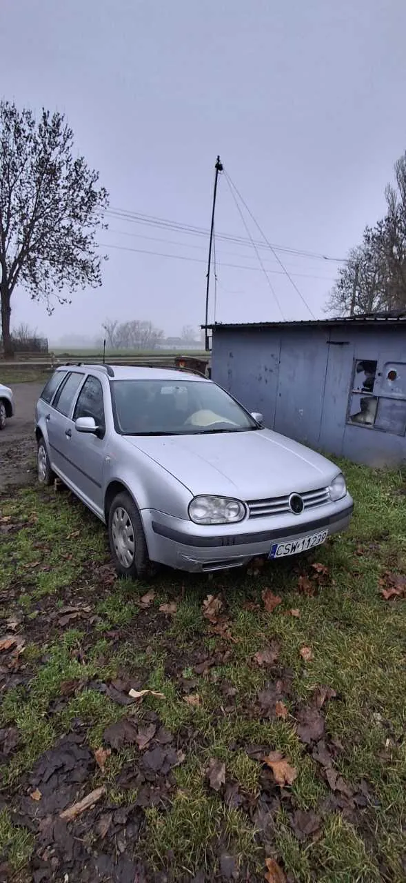 Srebrny samochód typu kombi zaparkowany na trawie z suchymi liśćmi. Widoczna tablica rejestracyjna CSW 11229. Po prawej stronie niebieski budynek gospodarczy z uszkodzonym oknem. W tle drzewa, linie energetyczne i mglisty krajobraz. Niebo jest pochmurne, na ziemi leżą suche liście.