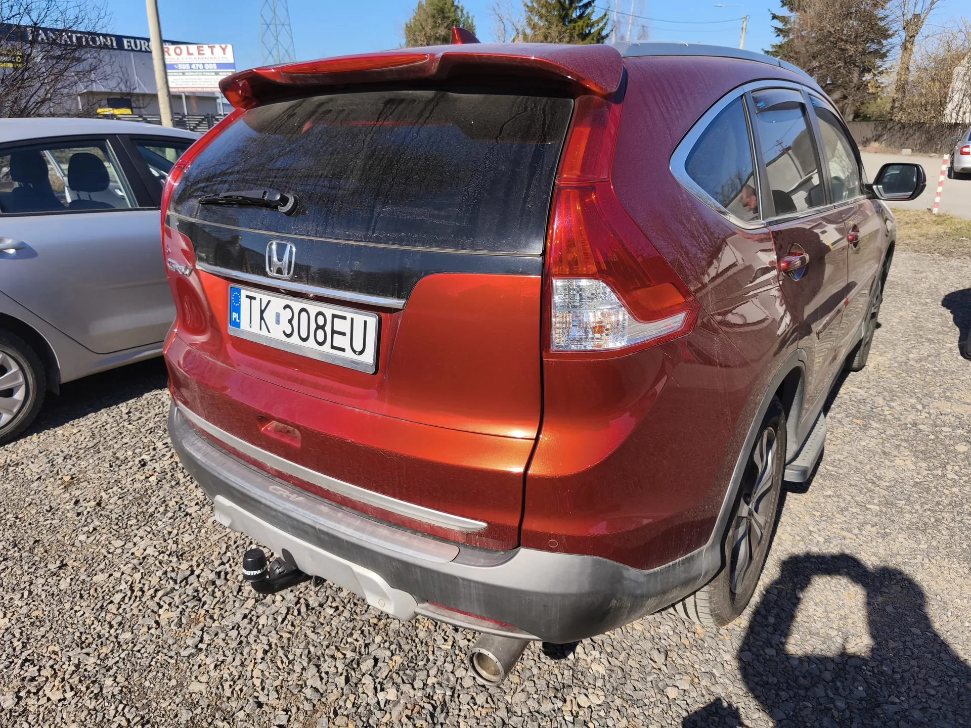 Czerwony samochód osobowy typu SUV marki Honda CR-V z tablicą rejestracyjną PL TK 308EU, zaparkowany na żwirowym placu. Widok z tyłu, z widocznym logo Hondy, tylnymi światłami i wydechami. W tle srebrny samochód, budynki z napisami "TANATONI EURO" i "ROLETY", drzewa oraz jasne niebo. Na ziemi widoczne cienie i drobny żwir.