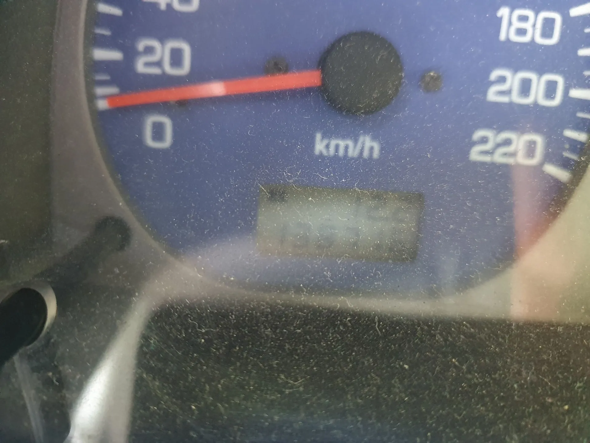 Zbliżenie na niebieski licznik prędkości w pojeździe. Tarcza zawiera białe cyfry: 0, 20, 180, 200 i 220 oraz oznaczenie jednostki 'km/h'. Czerwona wskazówka znajduje się w pozycji między 0 a 20. Poniżej znajduje się mały, prostokątny wyświetlacz cyfrowy, który jest nieczytelny. Powierzchnia licznika jest zakurzona.