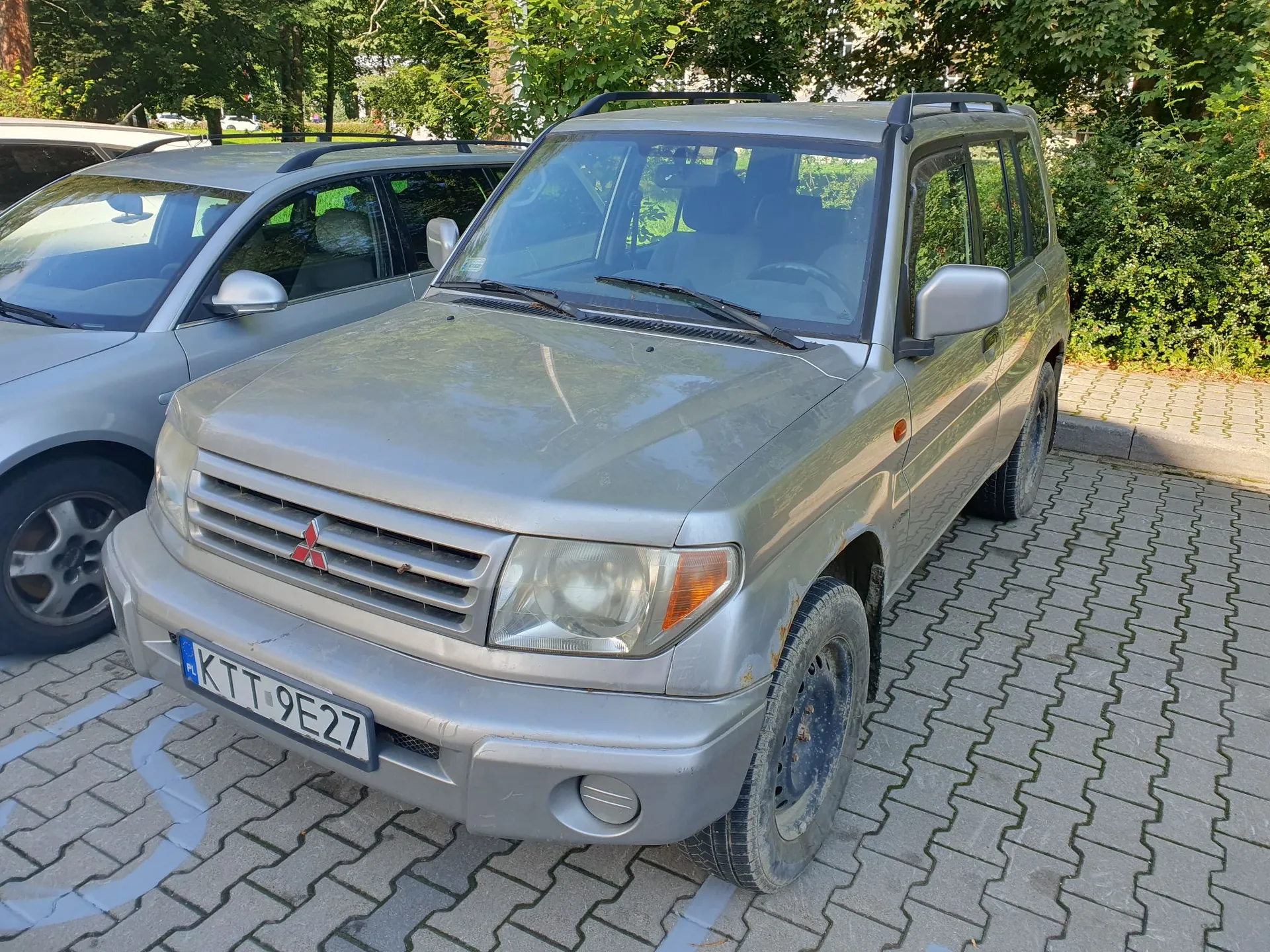 Srebrny samochód terenowy marki Mitsubishi zaparkowany na kostce brukowej. Pojazd ma widoczną tablicę rejestracyjną o numerze KTT 9E27. Obok niego stoi inny srebrny samochód. W tle widoczne są zielone drzewa i krzewy.