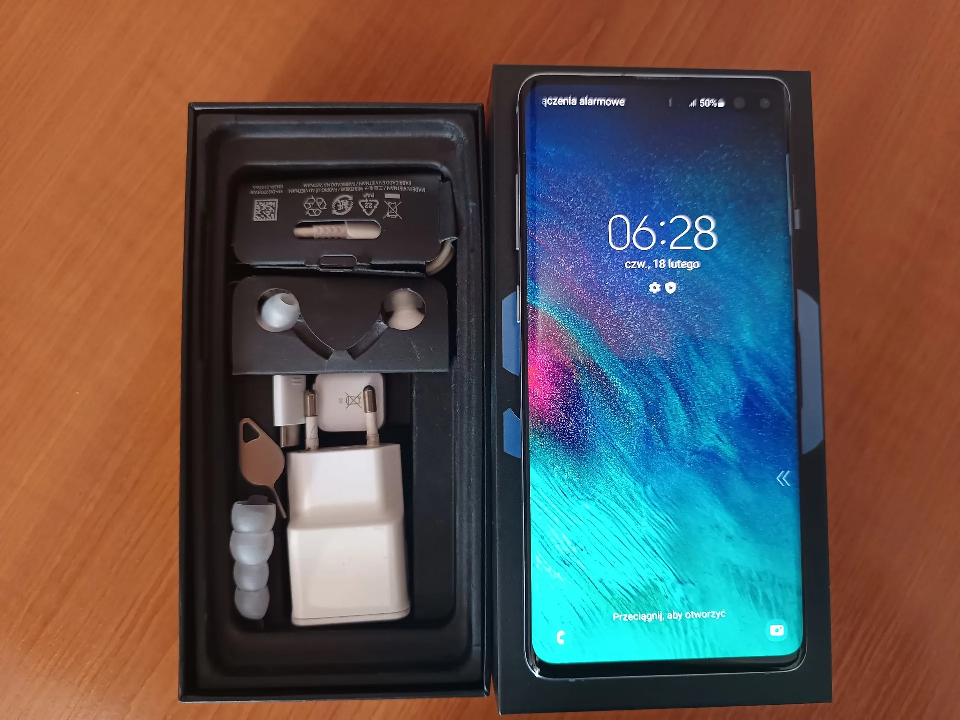 Zdjęcie przedstawiające otwarte czarne pudełko z zestawem akcesoriów do telefonu oraz włączony smartfon leżący obok. W pudełku znajdują się: biała ładowarka sieciowa, biały kabel USB, białe słuchawki douszne oraz zapasowe gumki do słuchawek. Ekran smartfona wyświetla godzinę 06:28, datę 'czw., 18 lutego' oraz napis na dole 'Przeciągnij, aby otworzyć'. W górnym pasku widoczny jest napis 'łączenia alarmowe' oraz poziom baterii 50%. Całość znajduje się na drewnianym blacie.
