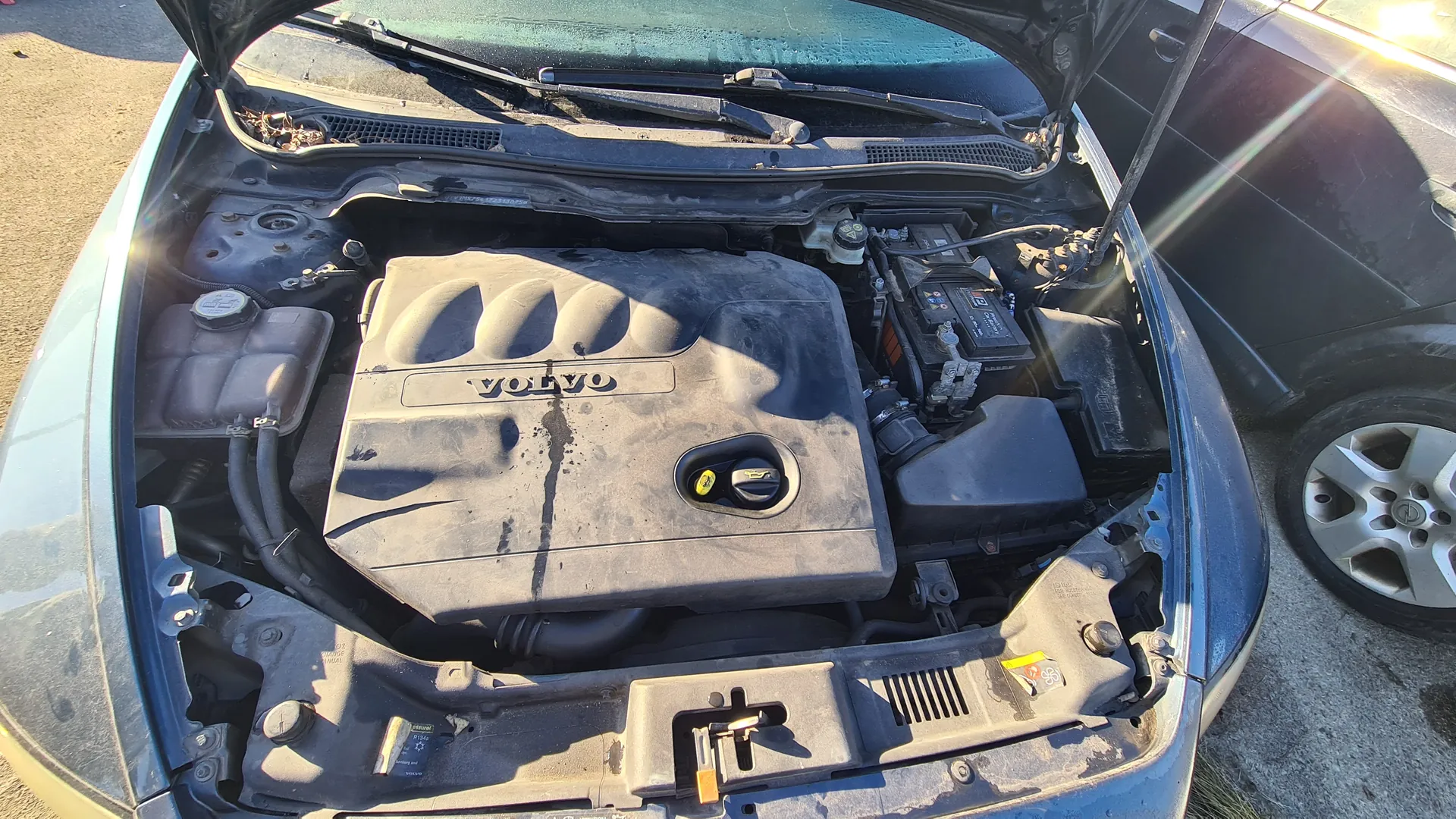 Widok otwartej maski samochodu marki Volvo z silnikiem. Na pokrywie silnika widoczny napis VOLVO. Silnik i elementy pod maską są zapylone. W prawej części obrazu widoczna część innego samochodu z kołem. Tło stanowi nawierzchnia asfaltowa. Słoneczne oświetlenie rzuca cienie na komponenty silnika.