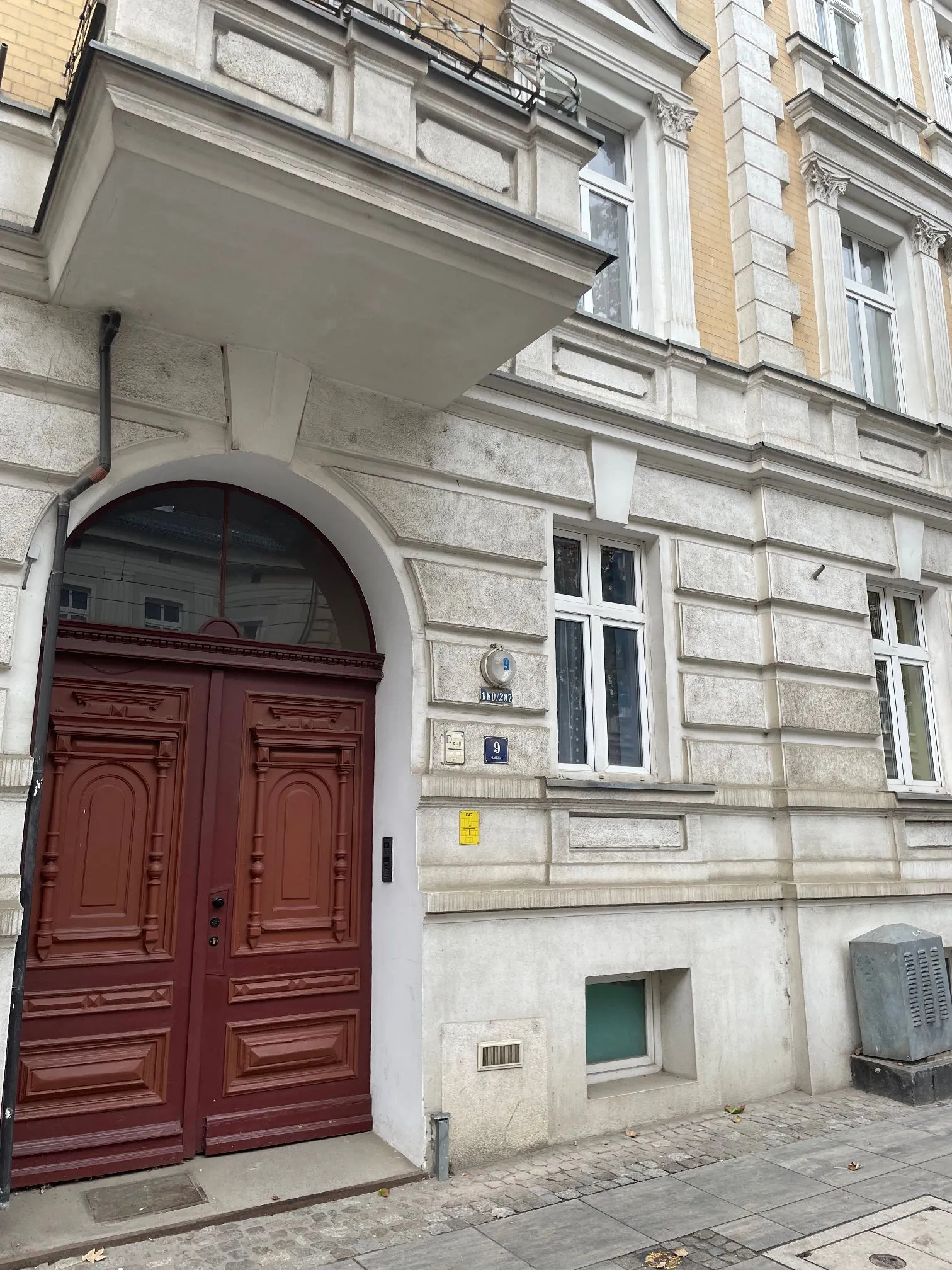 Zdjęcie fragmentu fasady jasnej, kamiennej kamienicy. Widoczne są duże, ciemnoczerwone, drewniane drzwi wejściowe z ozdobnymi rzeźbieniami i półkolistym nadprożem z przeszkleniem. Obok drzwi znajdują się tabliczki z numerem budynku '9' oraz inne mniejsze oznaczenia. W tle widać okna z białymi ramami oraz element wystający z budynku w górnej części obrazu. Budynek stoi przy chodniku ułożonym z szarych płyt i kostki brukowej.