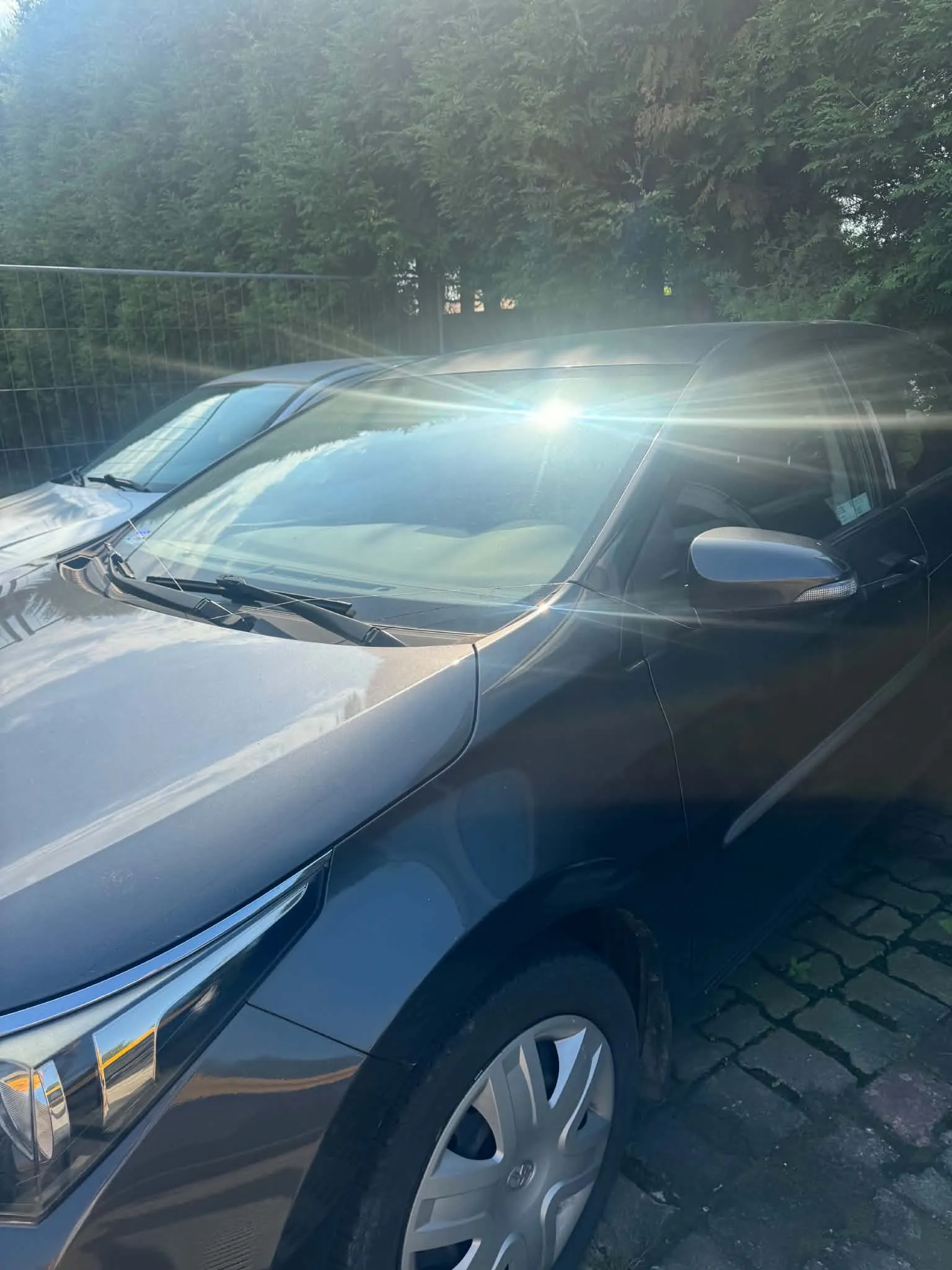 Zdjęcie przedstawiające przód i bok ciemnoszarego samochodu osobowego zaparkowanego na kostce brukowej. Auto jest oświetlone silnym słońcem, które odbija się od przedniej szyby i maski. W tle widoczny jest metalowy płot oraz gęste, zielone drzewa iglaste.