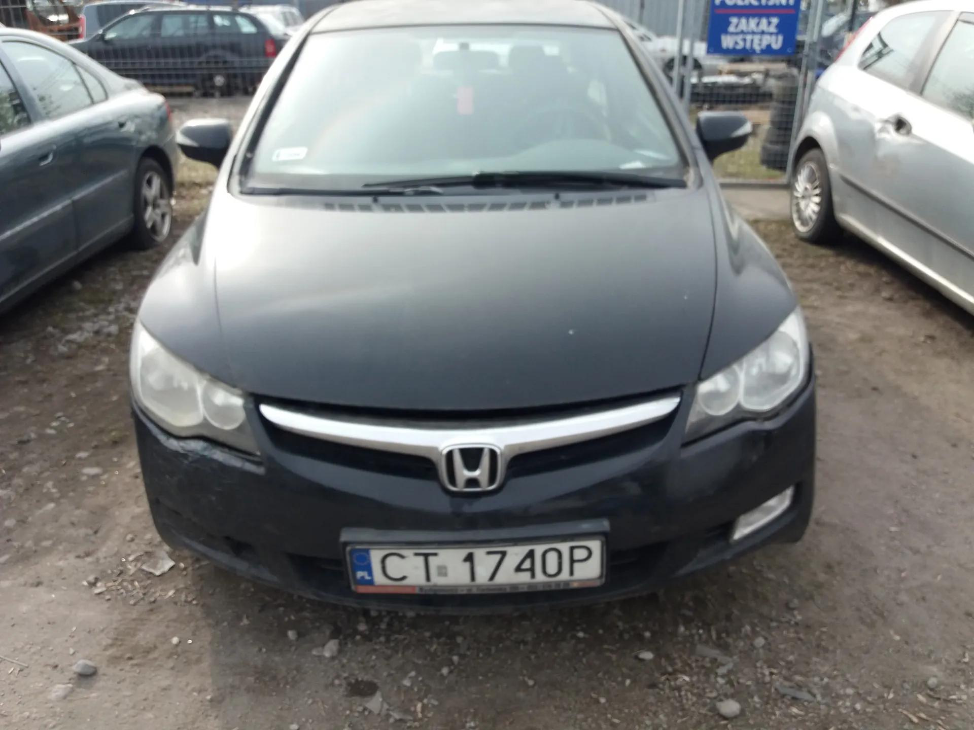 Czarny samochód marki Honda z tablicą rejestracyjną CT 1740P (PL) na pierwszym planie. W tle widoczne są inne zaparkowane pojazdy, ogrodzenie oraz niebieski znak z napisem ZAKAZ WSTĘPU. Podłoże stanowi żwirowa powierzchnia.