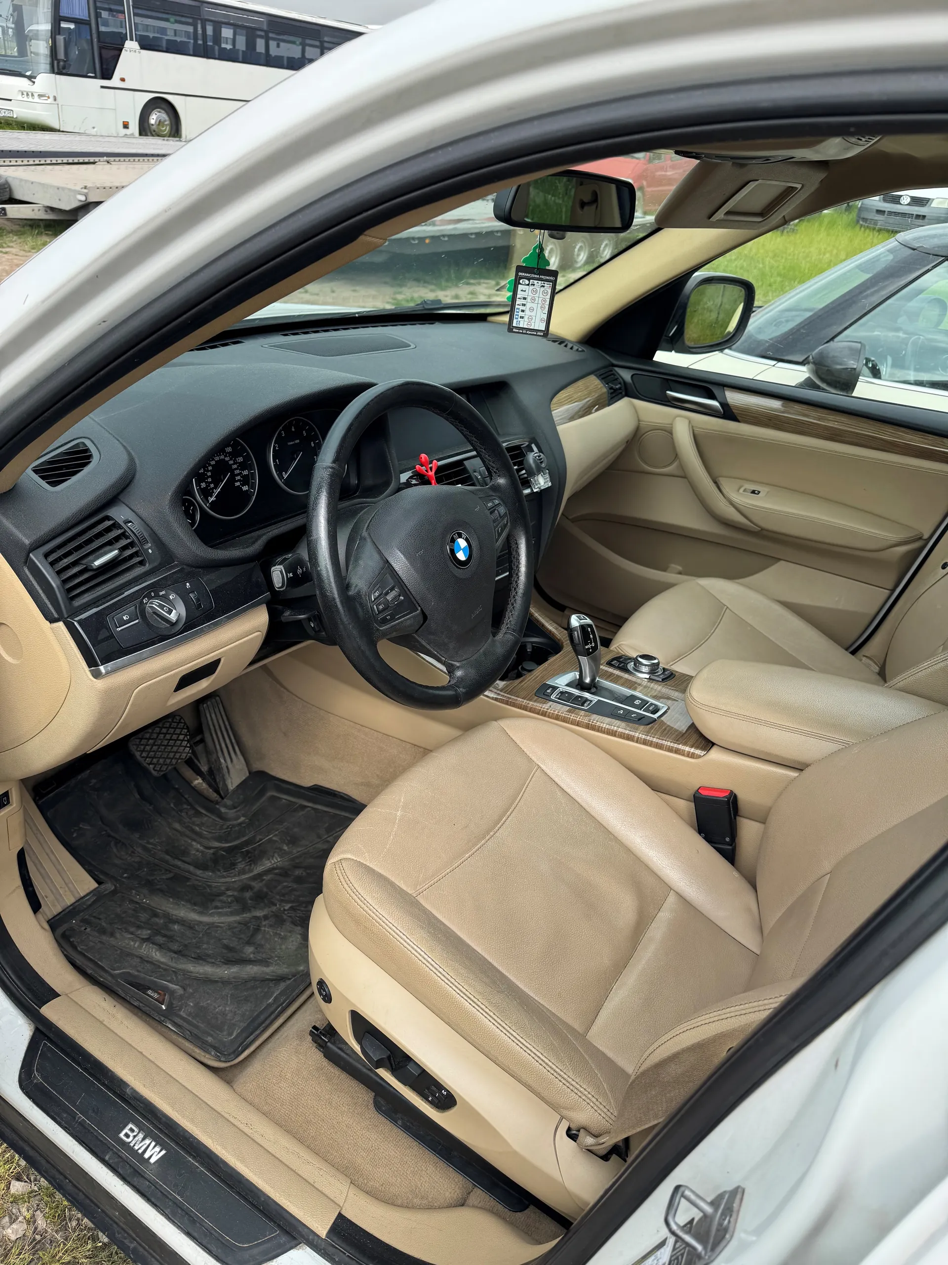 Wnętrze samochodu marki BMW z widocznym kierownicą z logo BMW, beżowymi skórzonymi fotelami, czarną deską rozdzielczą z dwoma okrągłymi wskaźnikami oraz centralną konsolą z przyciskami i dźwignią zmiany biegów. Na progu drzwi widoczny napis 'BMW'. W tle za przednią szybą widać autobus oraz inne pojazdy na zewnątrz. Wewnątrz samochodu umieszczono czarne dywaniki podłogowe, a na lusterku wstecznym wiszą ozdoby.
