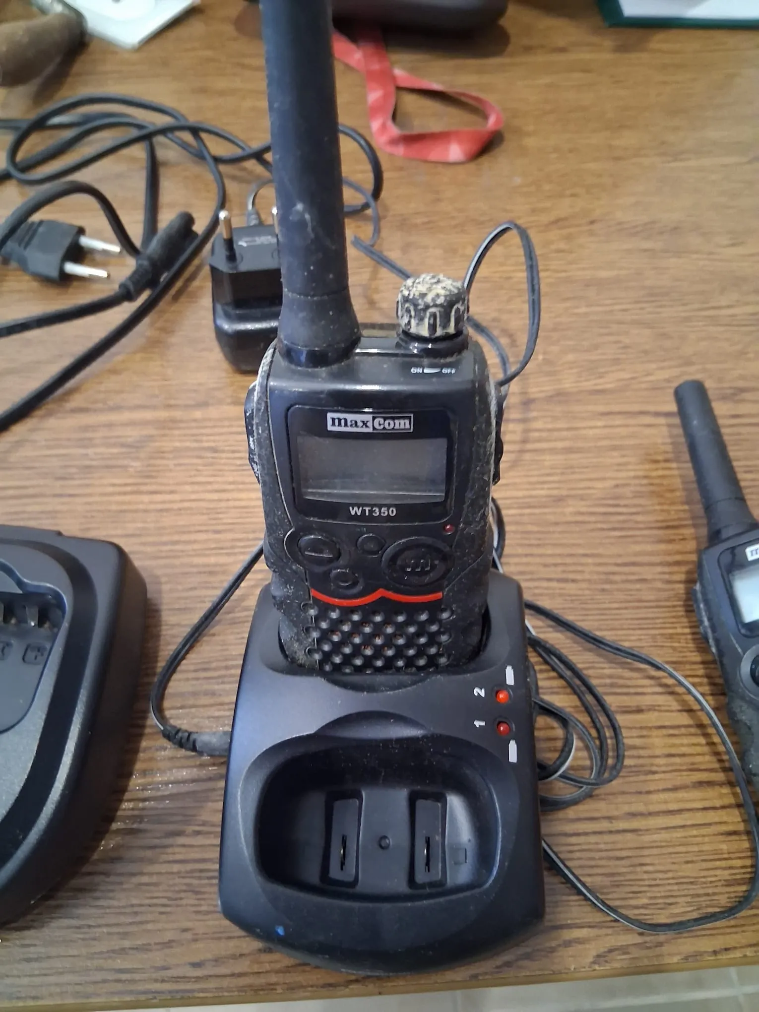 Czarny, zabrudzony radiotelefon marki maxCom, model WT350, umieszczony w stacji dokującej na drewnianym blacie. Urządzenie posiada długą antenę i przyciski sterujące. Obok znajdują się kable zasilające oraz fragment innego radiotelefonu.