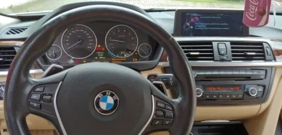 Widok z perspektywy kierowcy na wnętrze samochodu marki BMW. W centrum znajduje się czarna kierownica z logo BMW. Za nią widoczna jest deska rozdzielcza z analogowymi zegarami prędkości i obrotomierzem. Po prawej stronie znajduje się ekran multimedialny z wyświetlanym tekstem oraz panel sterowania klimatyzacją i radiem. Wnętrze wykończone jest w kolorze beżowym i czarnym.