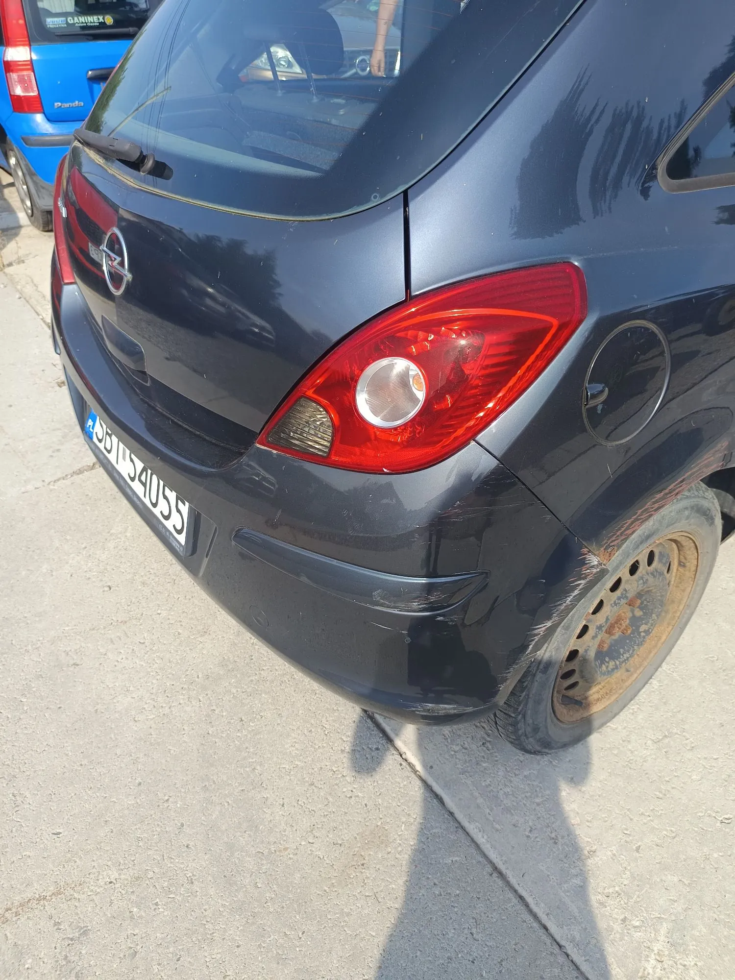 Zbliżenie na tył ciemnogranatowego samochodu z widocznym uszkodzeniem tylnego zderzaka po prawej stronie. Widoczna jest tylna lampa, logo marki Opel oraz fragment tablicy rejestracyjnej. W tle widać fragment niebieskiego samochodu marki Fiat Panda.