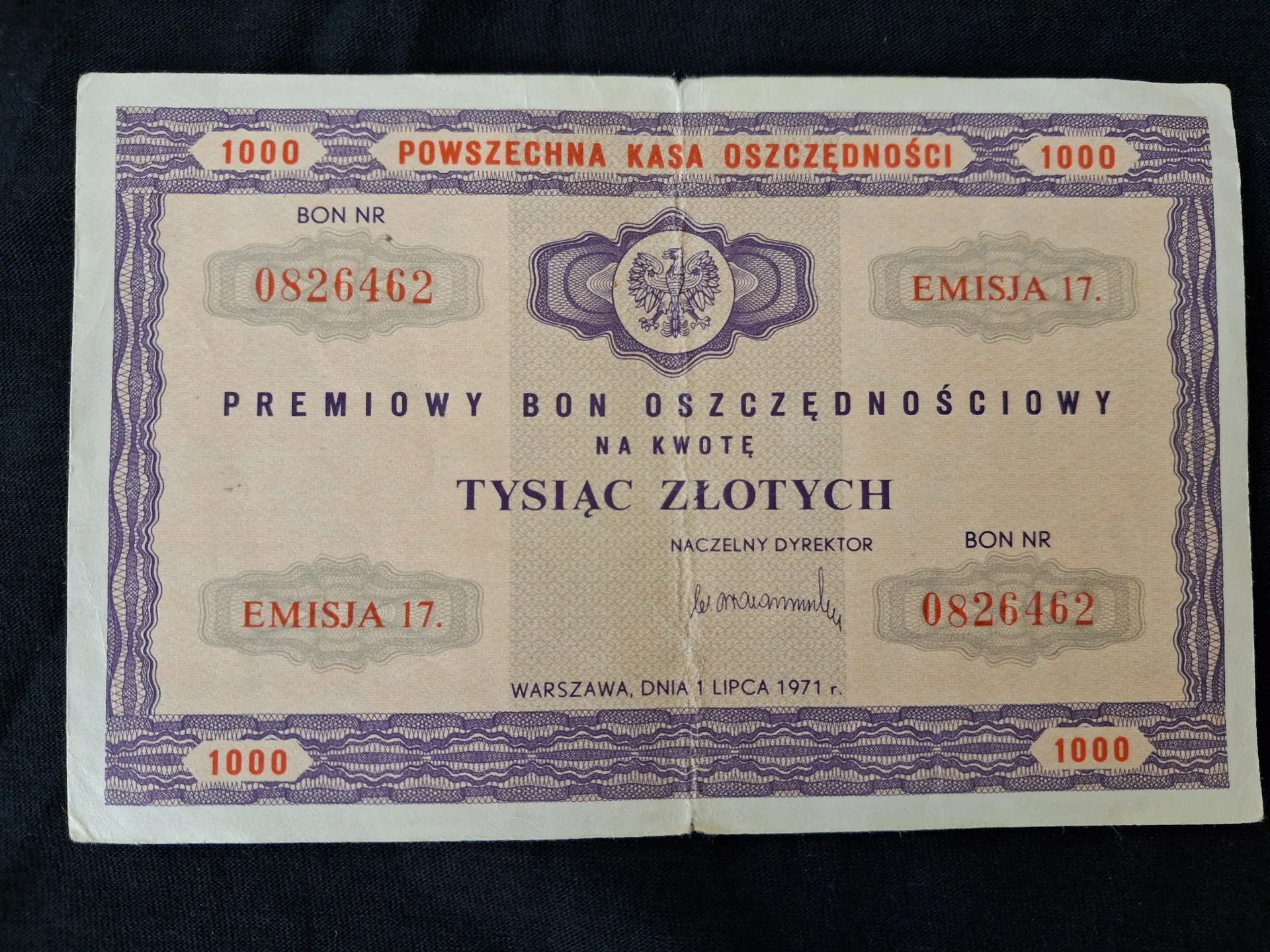 Zdjęcie starego, papierowego dokumentu o nazwie 'PREMIOWY BON OSZCZĘDNOŚCIOWY NA KWOTĘ TYSIĄC ZŁOTYCH'. Dokument wydany przez Powszechną Kasę Oszczędności w Warszawie dnia 1 lipca 1971 r. Na dokumencie widnieje numer bonu 0826462, oznaczenie 'EMISJA 17.' oraz godło Polski w centralnej części u góry. W dolnej części znajduje się podpis Naczelnego Dyrektora. Dokument ma fioletowe zdobienia ramkowe i powtórzone w rogach liczby 1000.