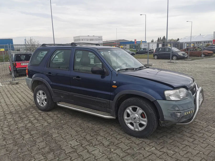 Ciemnoniebieski SUV z widocznymi uszkodzeniami na przednim zderzaku i bocznymi naczepami, zaparkowany na brukowanej powierzchni. W tle ogrodzenie, inne samochody (czerwony, czarny, brązowy), budynki przemysłowe, latarnie uliczne oraz pochmurne niebo.
