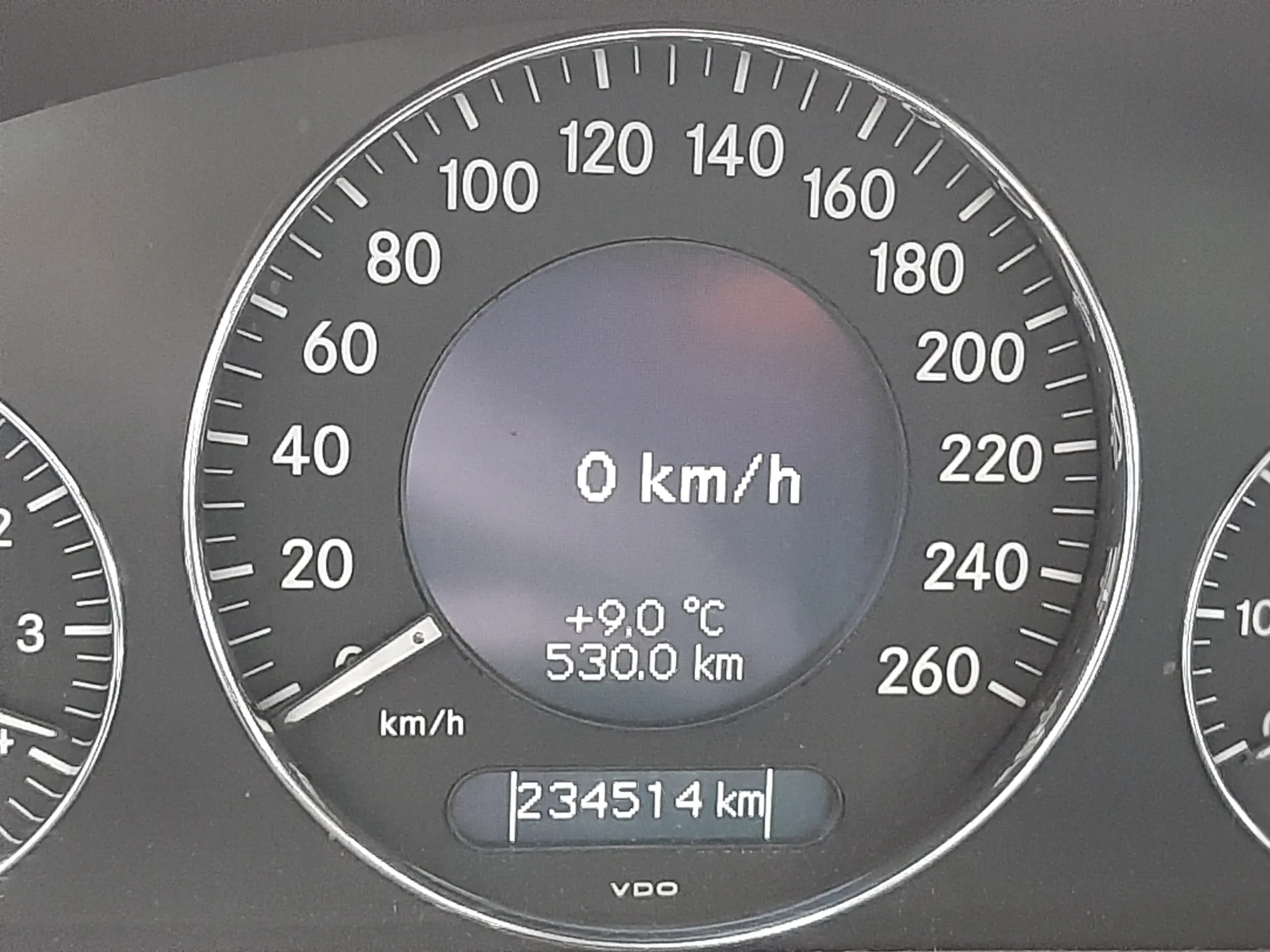 Zbliżenie na samochodowy wskaźnik prędkości z cyfrowym wyświetlaczem. Wskazówka prędkości pokazuje 0 km/h. W środku wyświetlacza widoczne są informacje: 0 km/h, +9,0 °C, 530,0 km. Poniżej znajduje się licznik przebiegu z cyframi 234514 km oraz napis VDO. Po lewej i prawej stronie widoczne są fragmenty innych wskaźników.