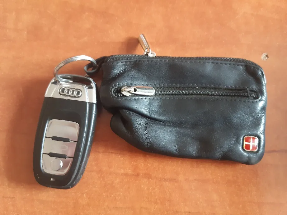 Czarny kluczyk do samochodu marki Audi z widocznym logo czterech pierścieni, przymocowany metalowym kółkiem do małego, czarnego, skórzanego portfela z zamkiem błyskawicznym. W prawym dolnym rogu portfela znajduje się mały, kwadratowy emblemat z biało-czerwonym krzyżem na czerwonym tle (flaga Danii). Przedmioty leżą na brązowej, drewnianej powierzchni.