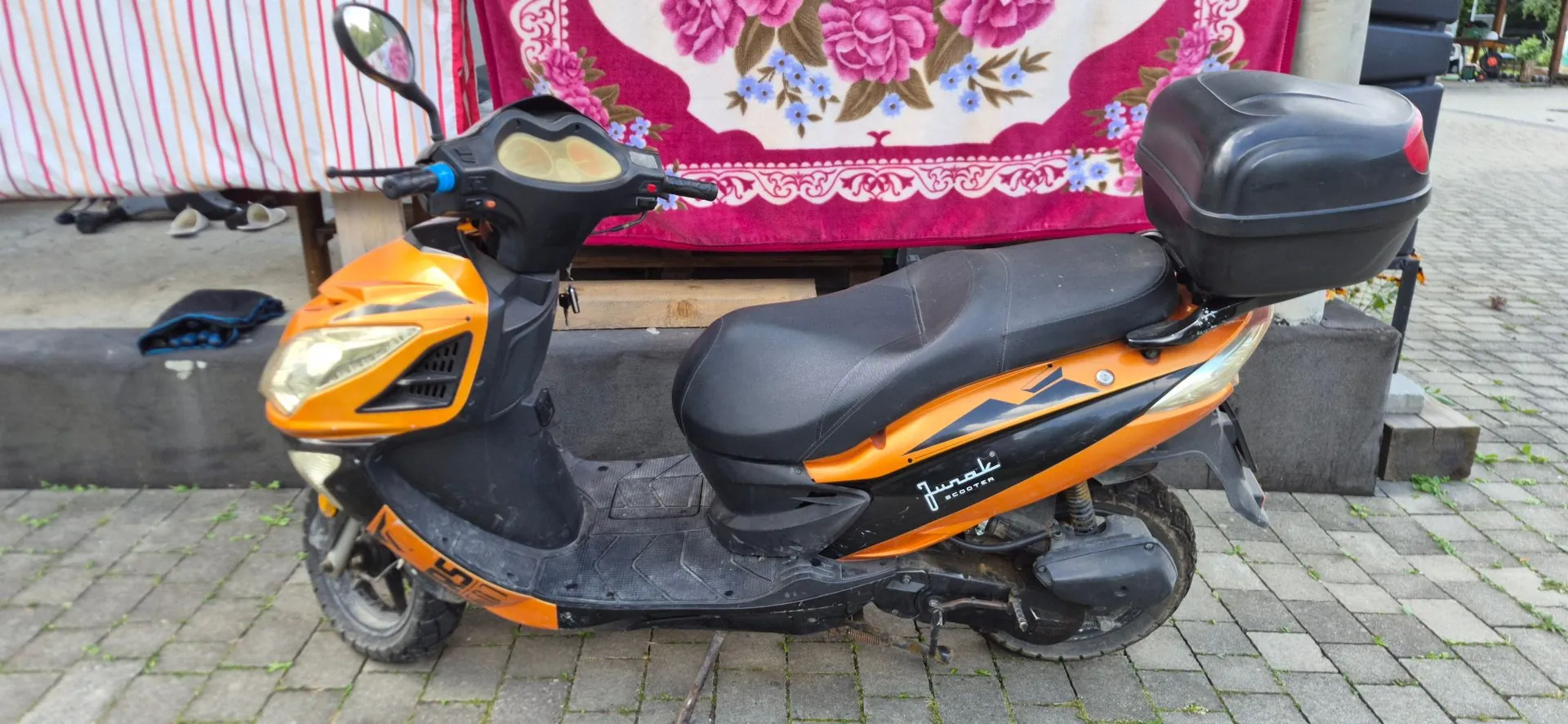 Pomarańczowo-czarny skuter marki Junak Scooter zaparkowany na kostce brukowej. Pojazd posiada czarną kanapę oraz czarny kufer zamontowany z tyłu. W tle widoczna jest czerwona tkanina z kwiatowym wzorem oraz szary krawężnik.