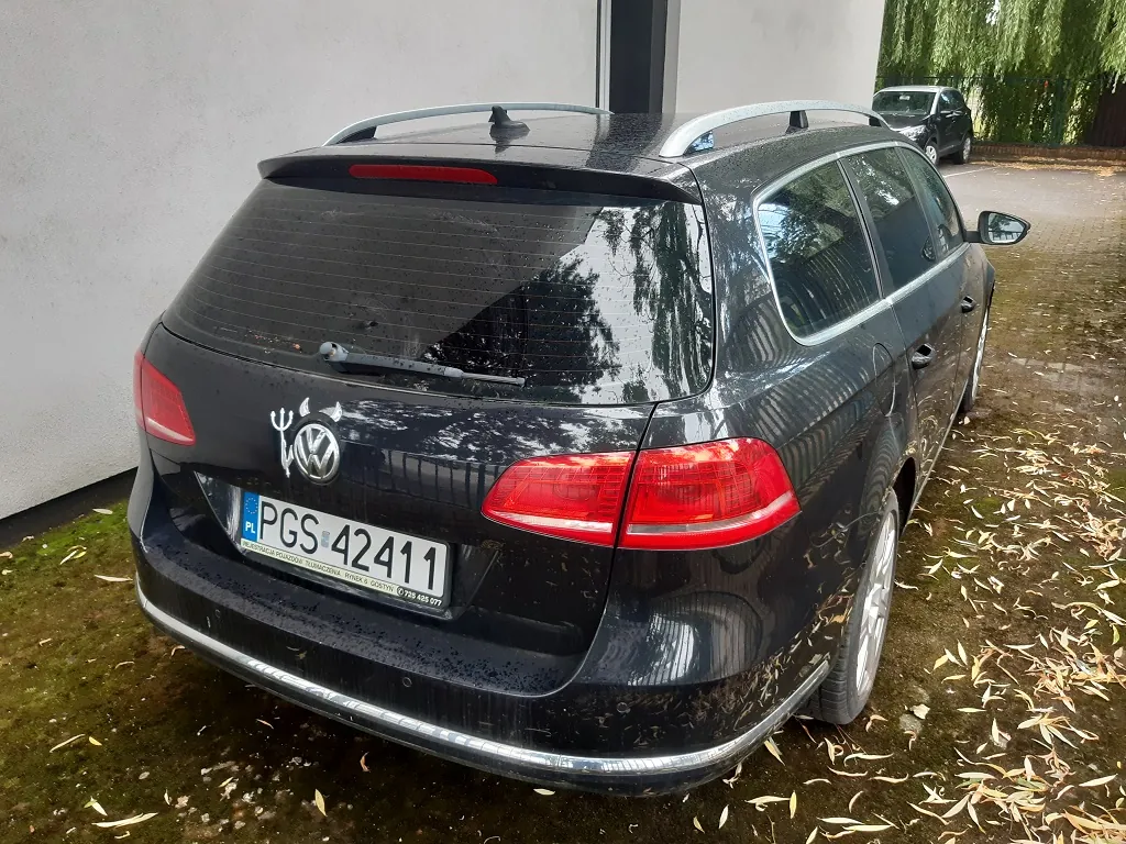 Tylny widok czarnego samochodu marki Volkswagen w nadwoziu kombi, zaparkowanego na podjeździe z mokrej kostki brukowej, na której leżą żółte liście. Samochód ma polską tablicę rejestracyjną o numerze PGS 42411. Na klapie bagażnika, obok logo Volkswagena, znajduje się naklejka przedstawiająca sylwetkę byka z widelcem trójzębem. W tle widoczny jest biały budynek oraz inny ciemny samochód zaparkowany dalej.