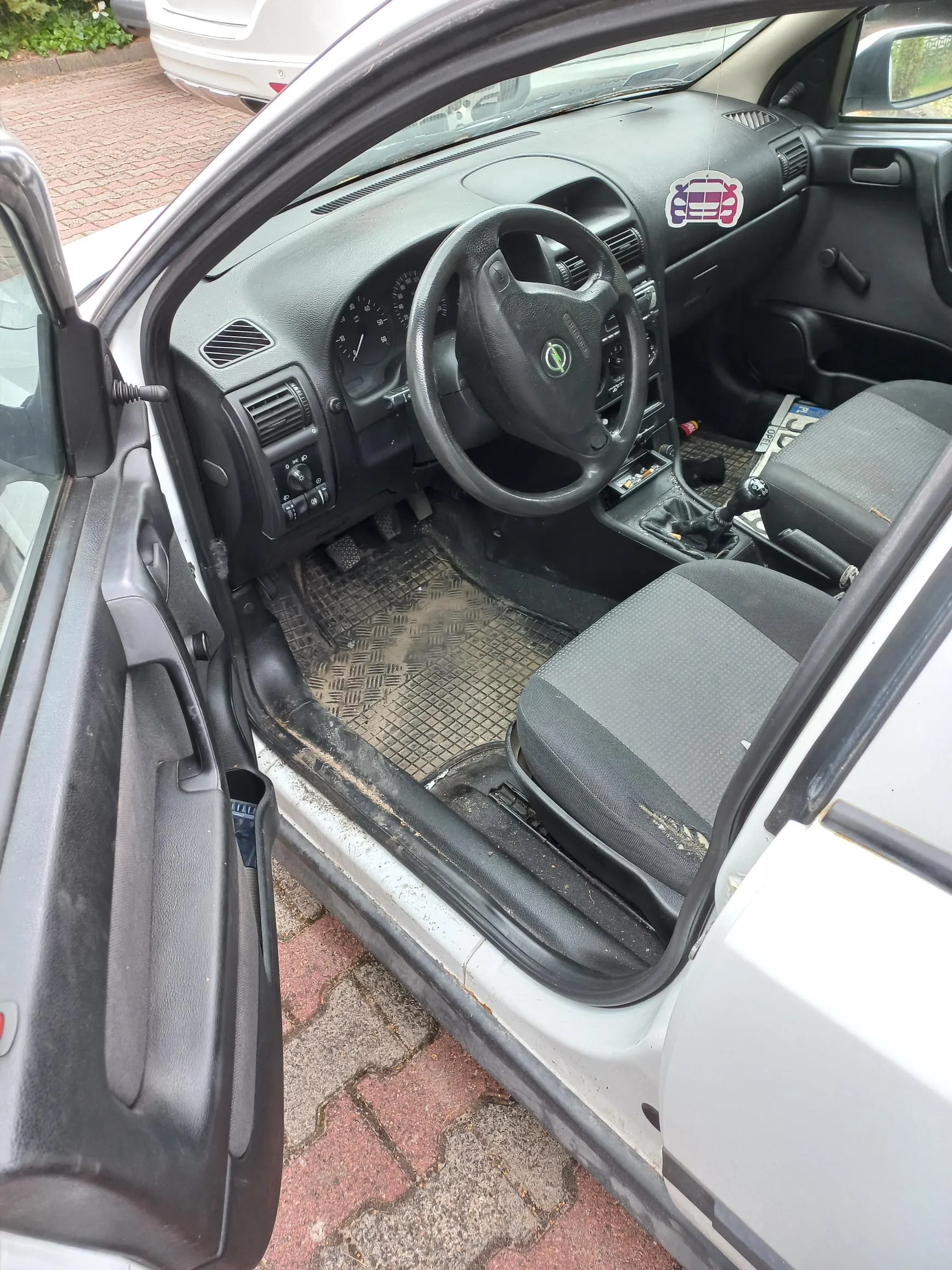 Widok z otwartych drzwi kierowcy do wnętrza srebrnego samochodu marki Opel. Widoczna jest czarna kierownica z logo producenta, deska rozdzielcza z zegarami, lewarek zmiany biegów oraz brudna, gumowa mata na podłodze. Na desce rozdzielczej znajduje się naklejka z czerwonym samochodem. Fotel kierowcy jest obity ciemnoszarą tkaniną.