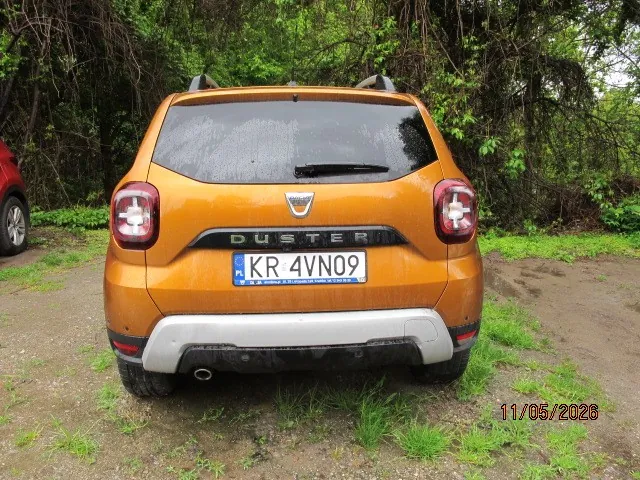 Z tyłu pomarańczowy samochód Dacia Duster z tablicą rejestracyjną KR 4VN09. W tle zielone krzewy i drzewa, podłoga z drobnej ziemi i trawy. W prawym dolnym rogu widoczna data 11/05/2026. Po lewej stronie fragment czerwonego pojazdu.