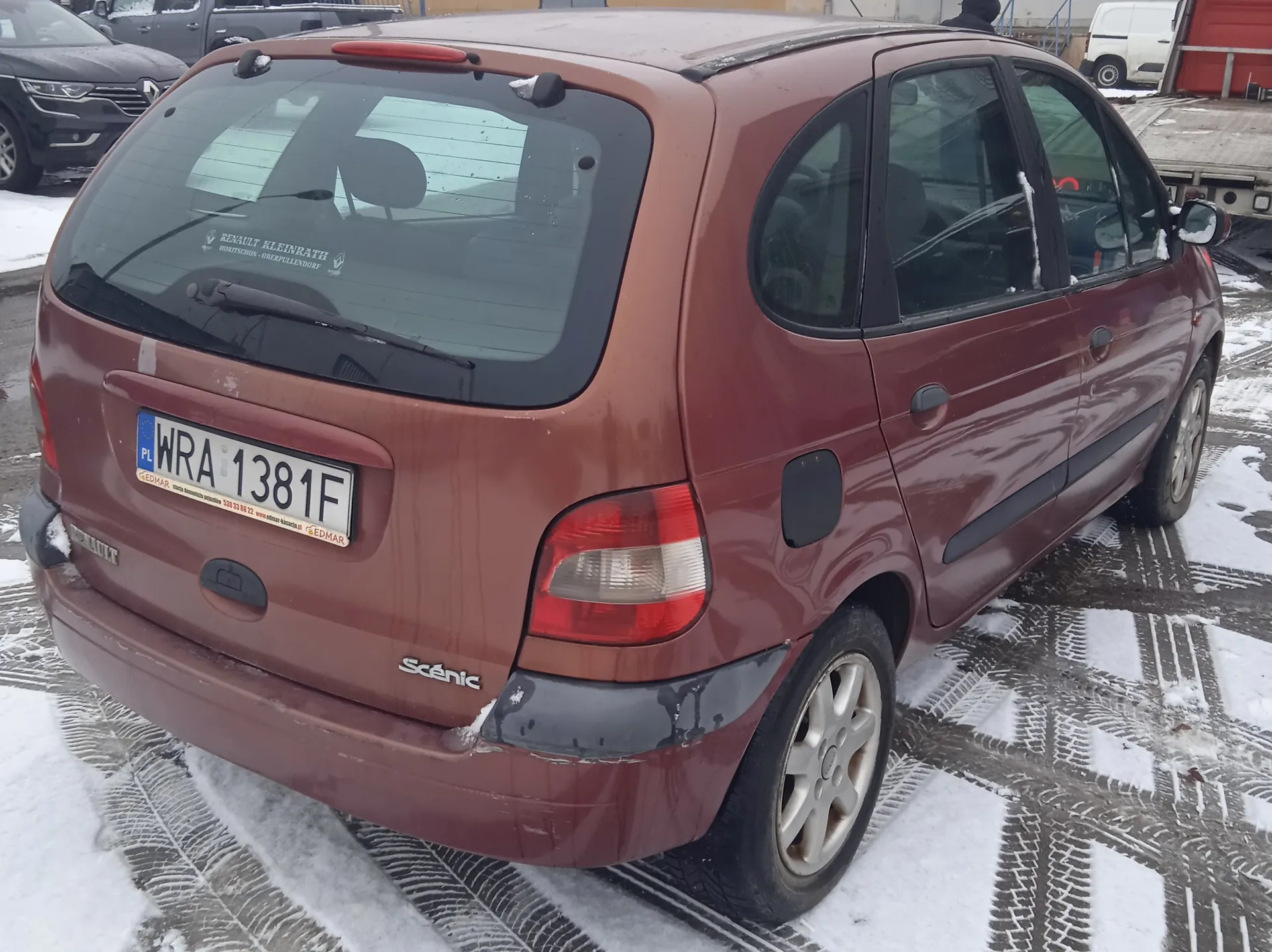 Czerwony samochód Renault Scénic zaparkowany na śnieżnej nawierzchni z odciskami opon. Widoczny tył pojazdu z tablicą rejestracyjną WRA 1381F oraz napisem "Scénic" na bagażniku. Na szybie tylnej znajduje się napis "RENAULT KLEINRATH". W tle inne pojazdy, w tym czarny samochód i biały furgon, oraz budynki. Śnieg pokrywa część podwozia i koła.