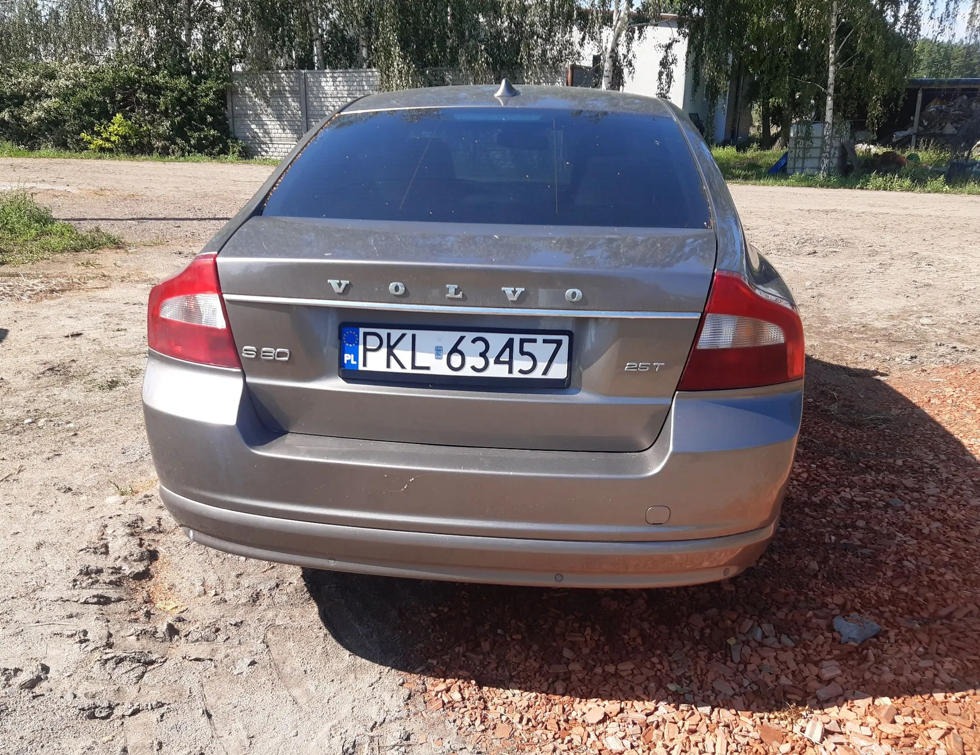 Tylny widok szarego samochodu marki Volvo S60 2.5T. Pojazd posiada polską tablicę rejestracyjną o numerze PKL 63457. Samochód stoi na piaszczystym podłożu z fragmentami czerwonej kruszywy ceglanej. W tle widoczne są drzewa i jasny płot.