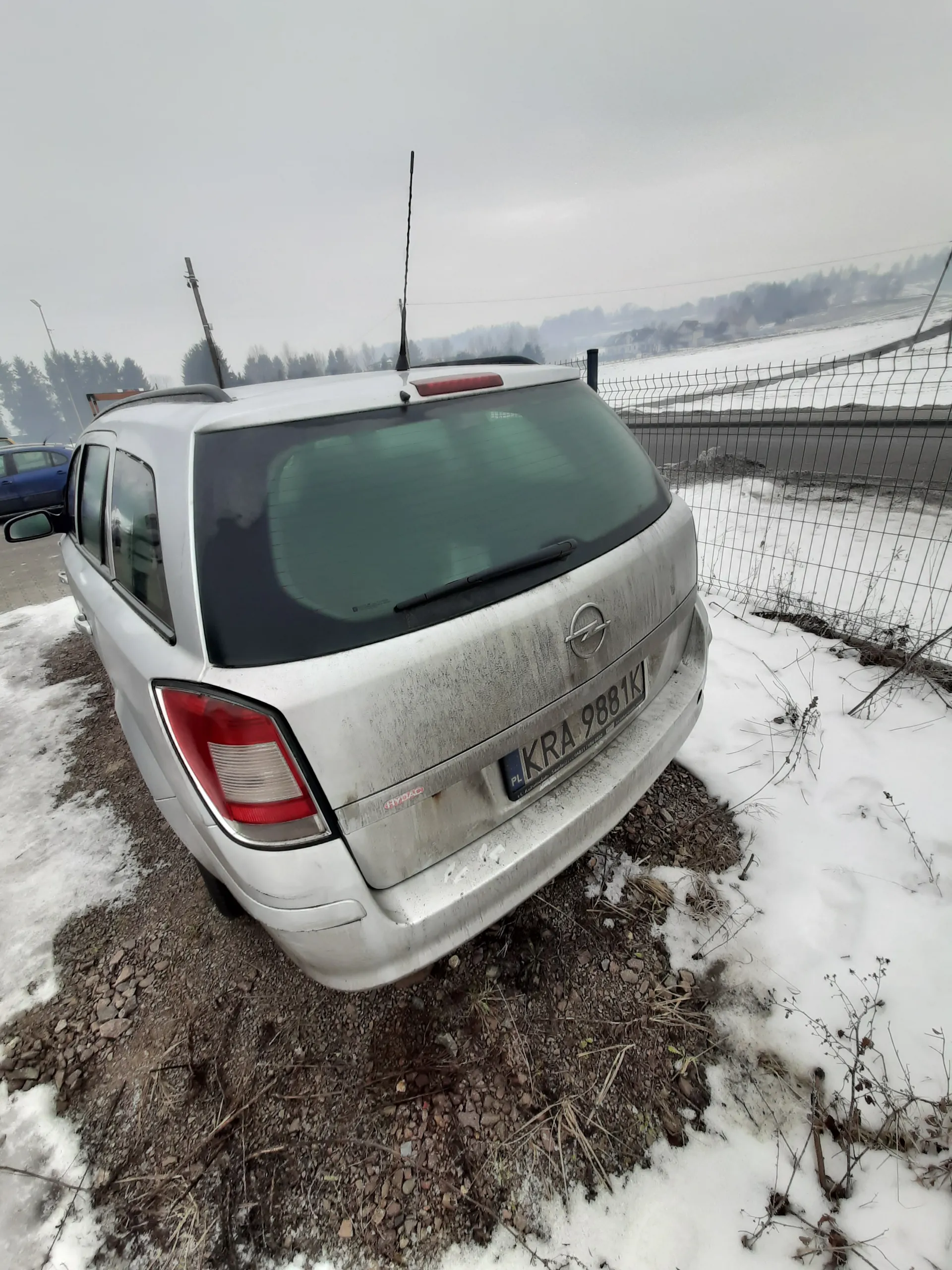Tył srebrnego samochodu z tablicą rejestracyjną KRA 9881K umieszczoną na bagażniku. Samochód stoi na żwirowym podłożu z resztkami śniegu. W tle widoczna siatka ogrodzeniowa, droga oraz zalesiony teren. Niebo jest pochmurne. Samochód jest zabrudzony, a na bagażniku znajduje się logo marki.